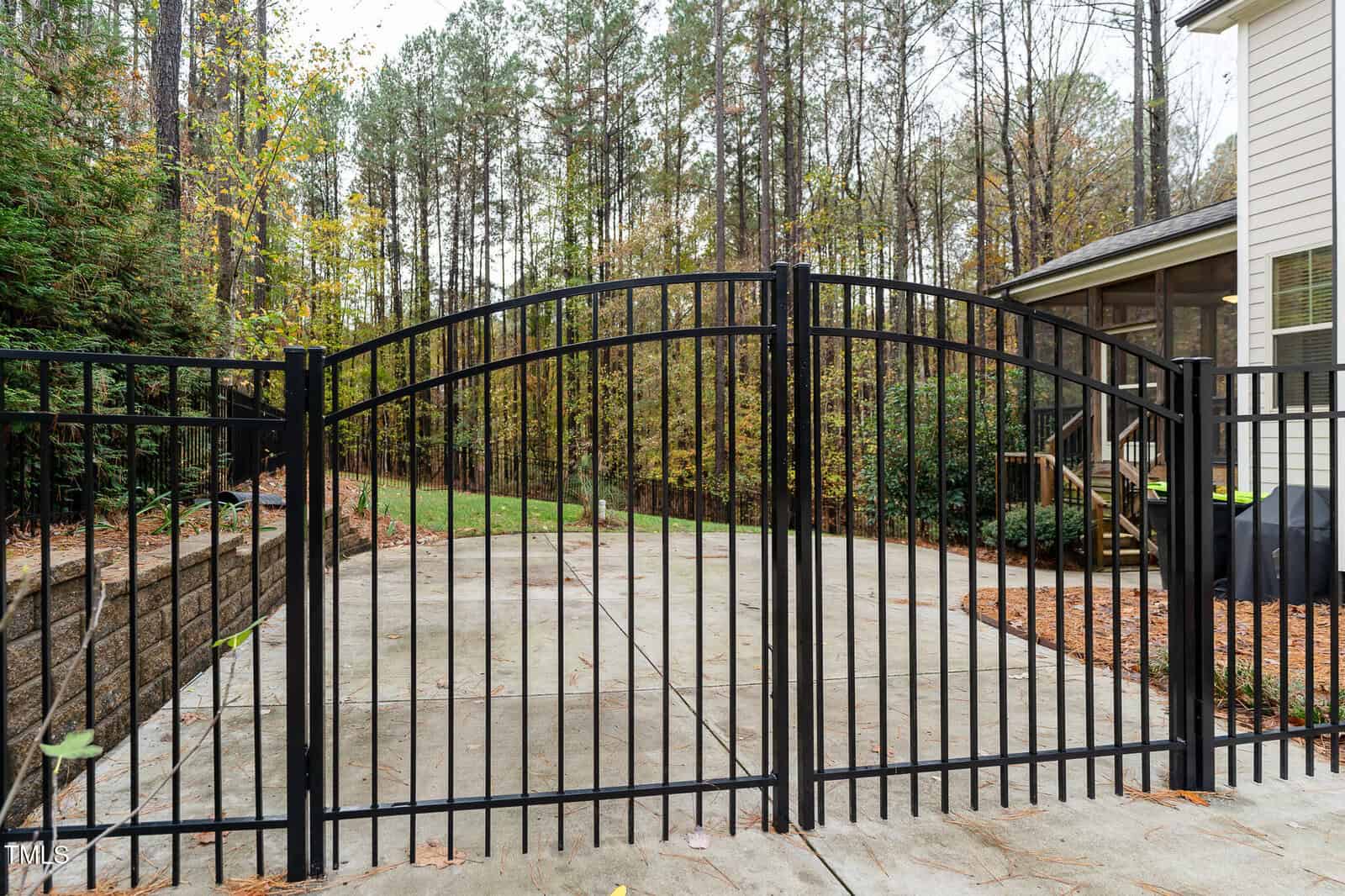 A black metal gate with curved tops encloses a concrete driveway. Trees and dense vegetation are visible in the background, and the corner of a building with windows and a small porch is on the right side of the image.