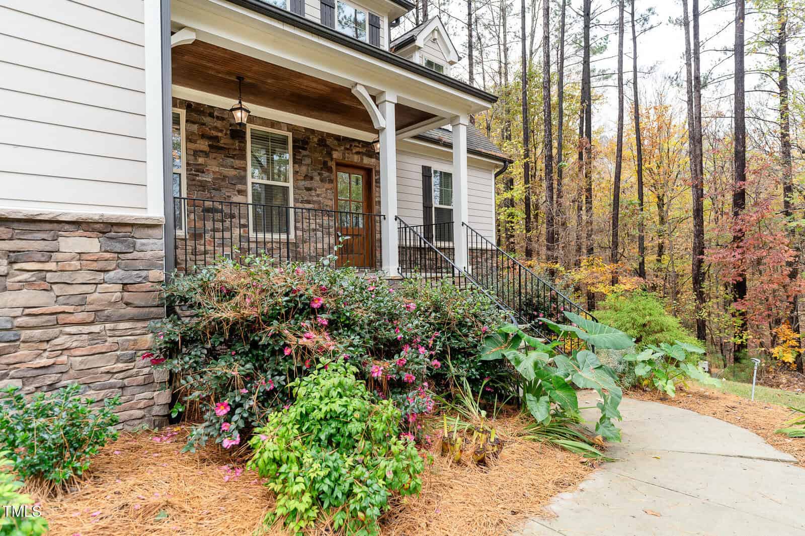 Front view of a house with a stone facade and a wooden door. A small front porch is bordered by black metal railings. The garden features various shrubs and blooming flowers, with a background of tall trees showing autumn colors.