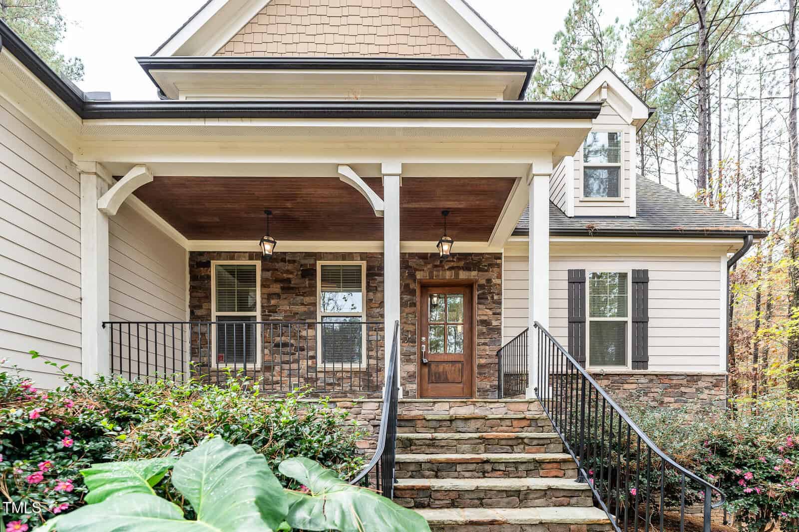 A house with a stone and shingle facade features a covered porch with two lantern-style lights. The entrance has a wooden door with a small window. Stone steps lead up to the porch, surrounded by shrubs and a few large plants.