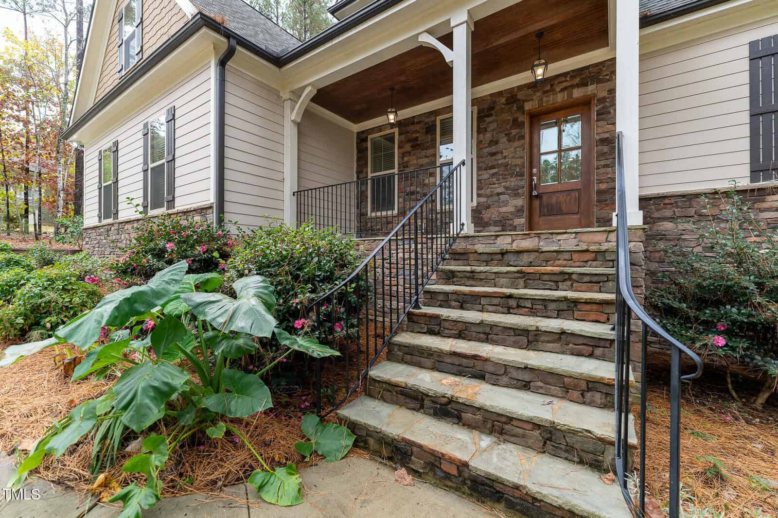 A house with stone accents features a wooden front door and a set of stone steps leading up to a covered porch. Large leafy plants and pink flowers grow beside the steps, and the house is surrounded by trees.