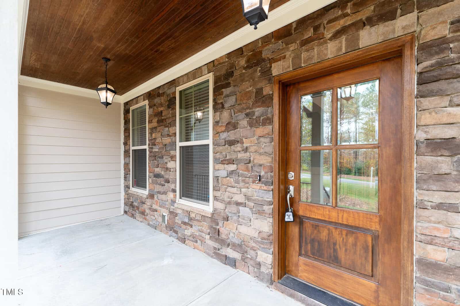 A porch with a wooden door featuring a glass panel and a lockbox. The exterior wall is made of stone, and there are two large windows. The ceiling is wood-paneled with a hanging light fixture. The floor is concrete.