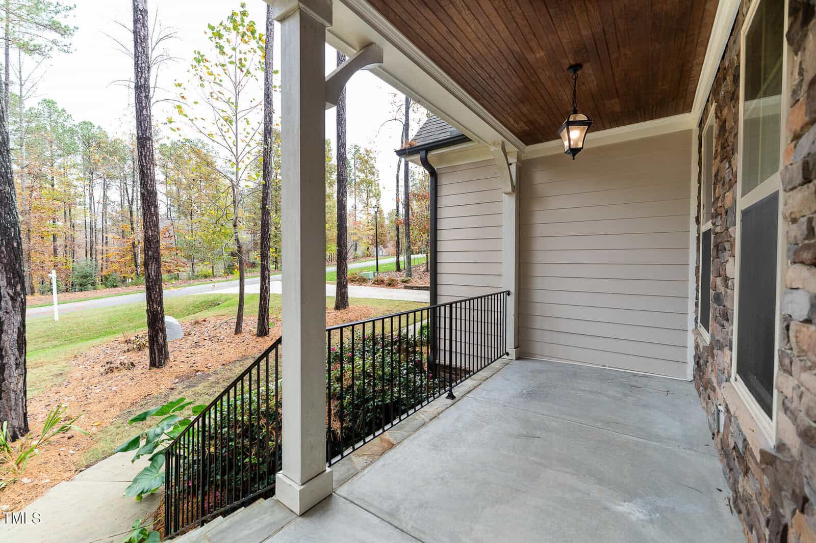 A covered porch with a wooden ceiling and a single hanging light fixture. The porch has a stone column and is adjacent to a railing. The view includes trees and a road in the background.