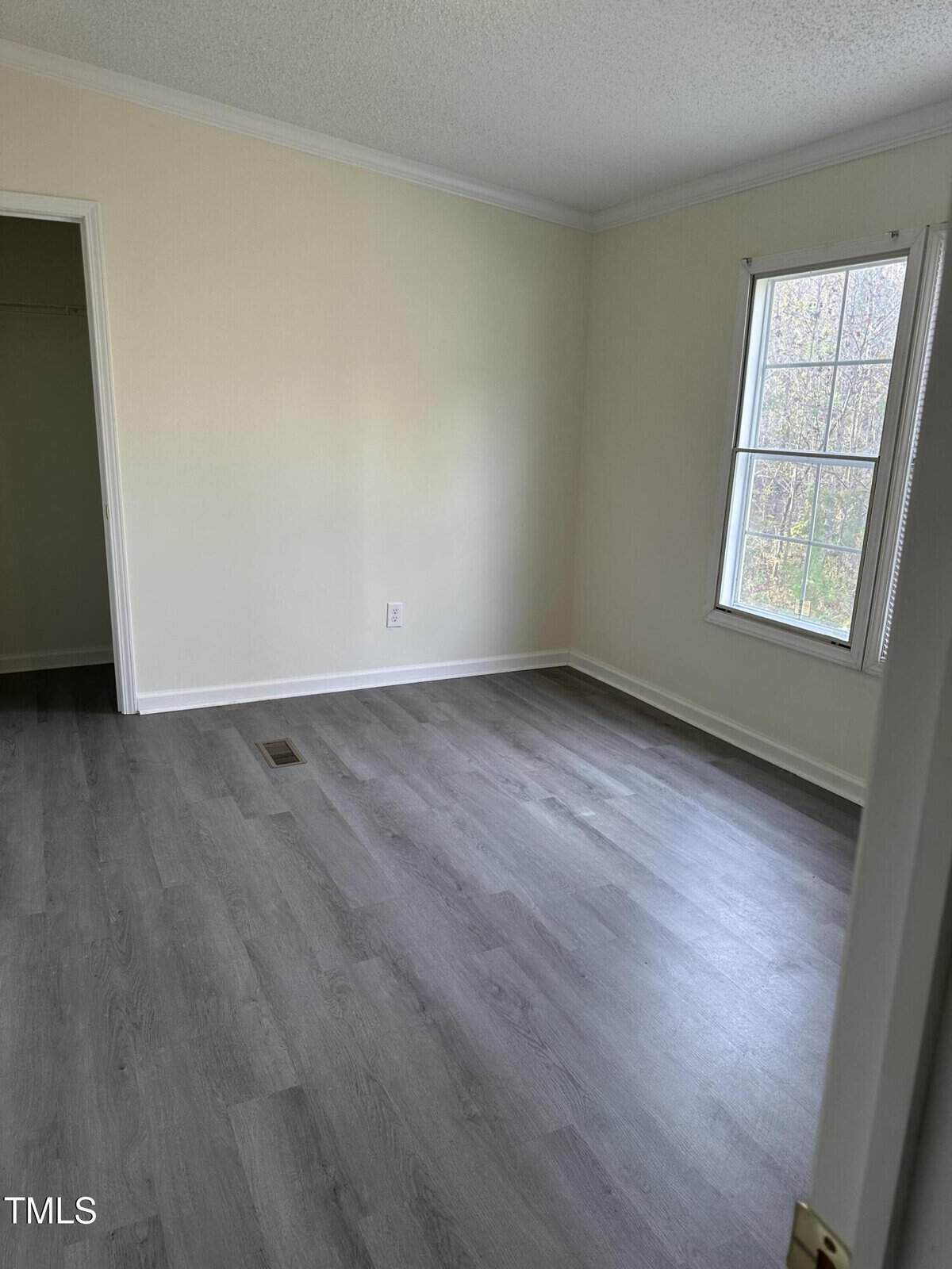Empty room with light-colored walls and hardwood flooring. A window on the right lets in natural light. An open door on the left leads to a small closet.