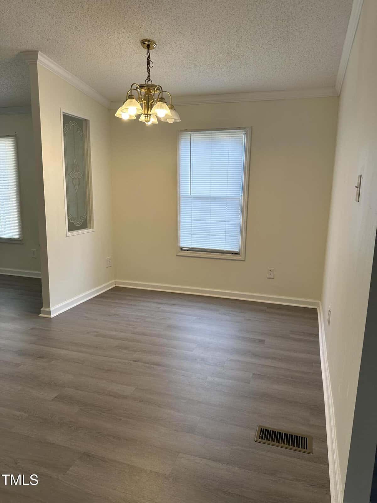 Empty room with light-colored walls and wood flooring. A window with blinds lets in natural light. A brass chandelier hangs from the ceiling, and a small decorative glass panel is set in a dividing wall.