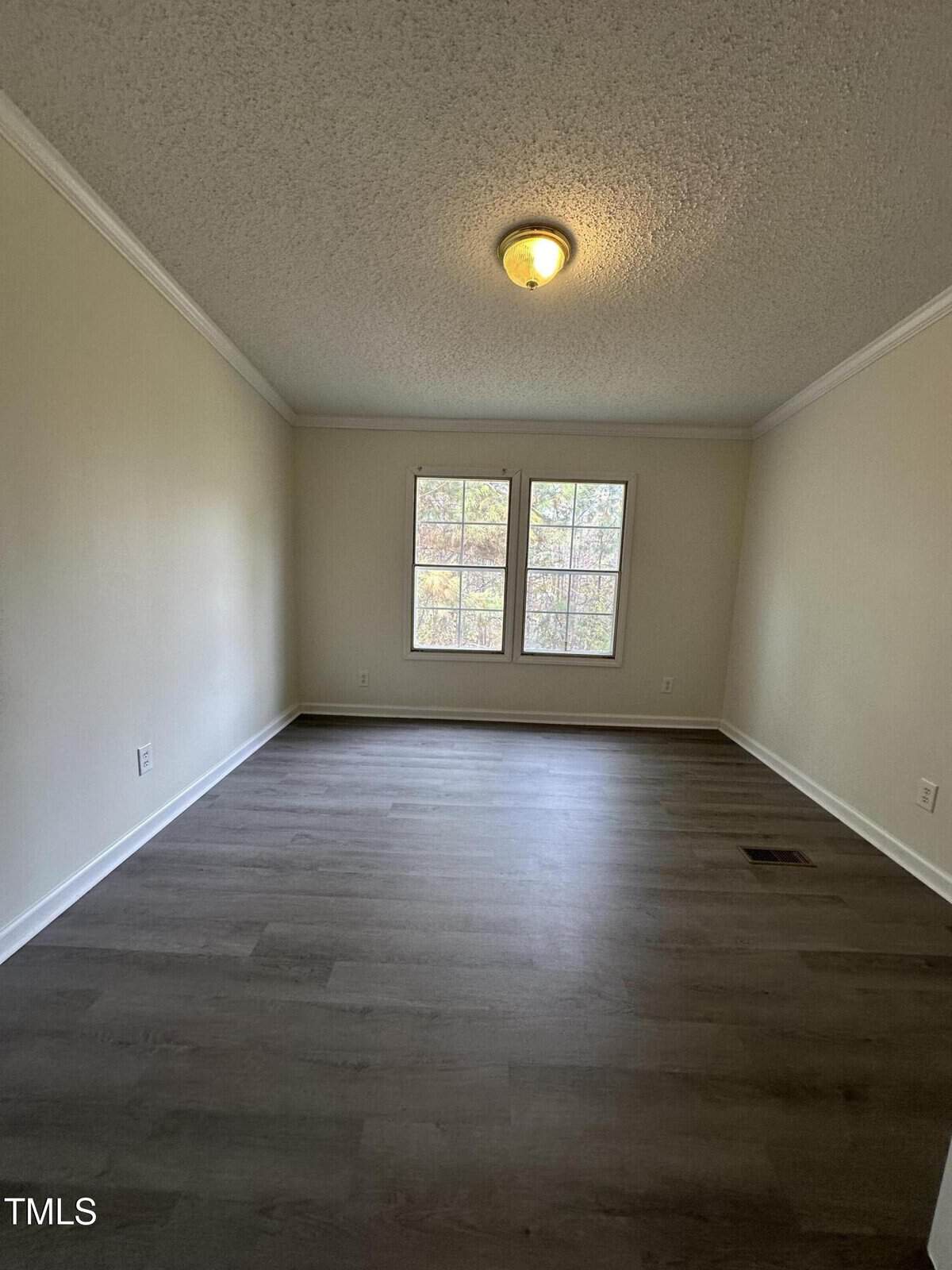 An empty room with light-colored walls and dark wooden flooring. A ceiling light fixture is centered, and two windows allow natural light to enter.