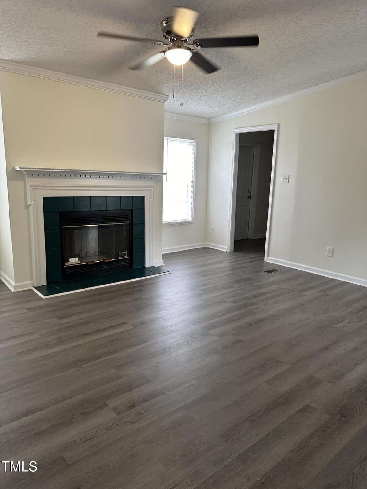 A living room with light gray hardwood floors, off-white walls, and a ceiling fan with lights. A fireplace with a dark green tile surround is centered on the left wall. A door and a large window are visible on the right side.