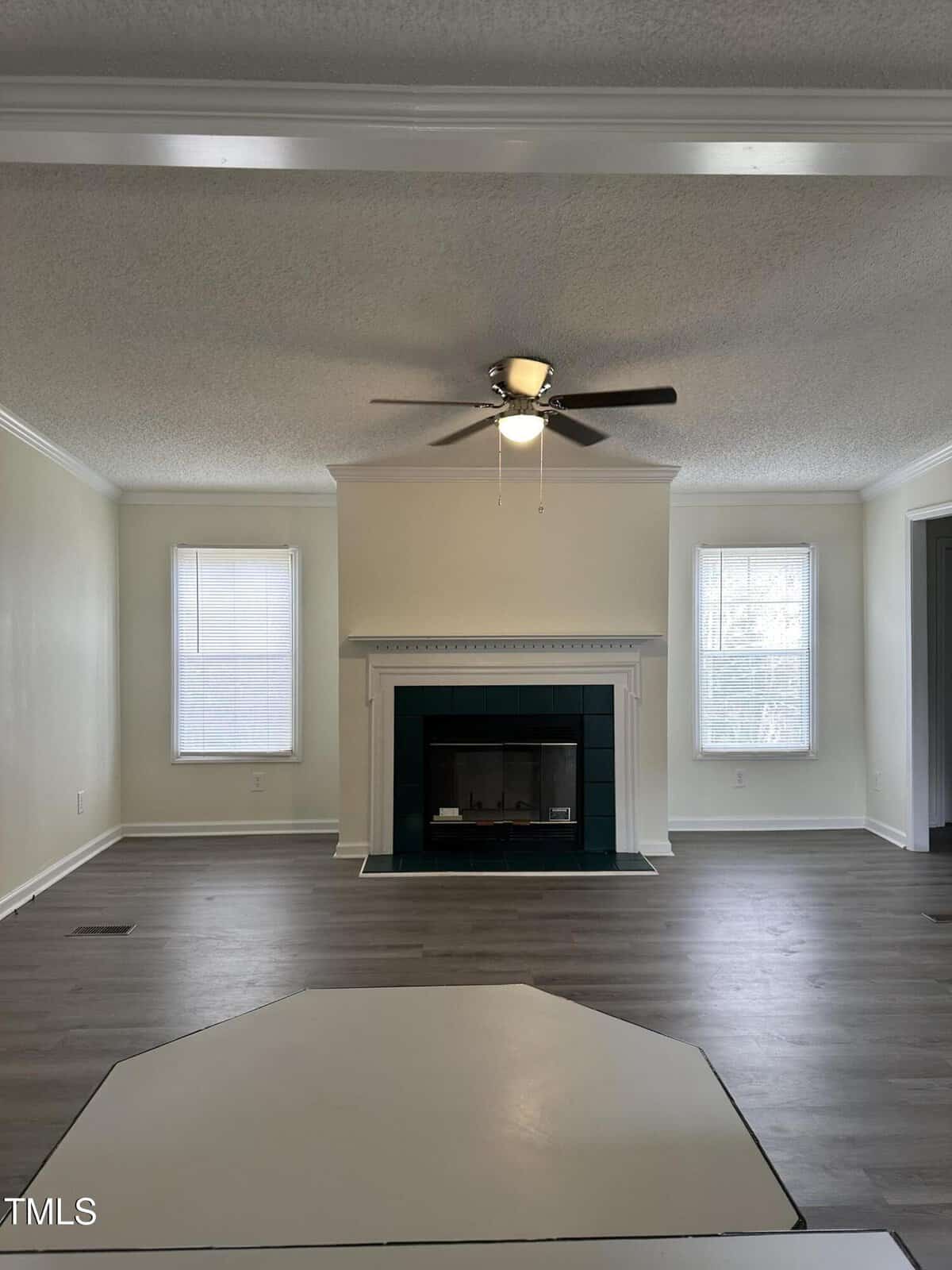 A living room with light-colored walls and wood flooring features a central fireplace framed by two tall windows. A ceiling fan with a light is mounted on the ceiling. The room appears spacious and empty.