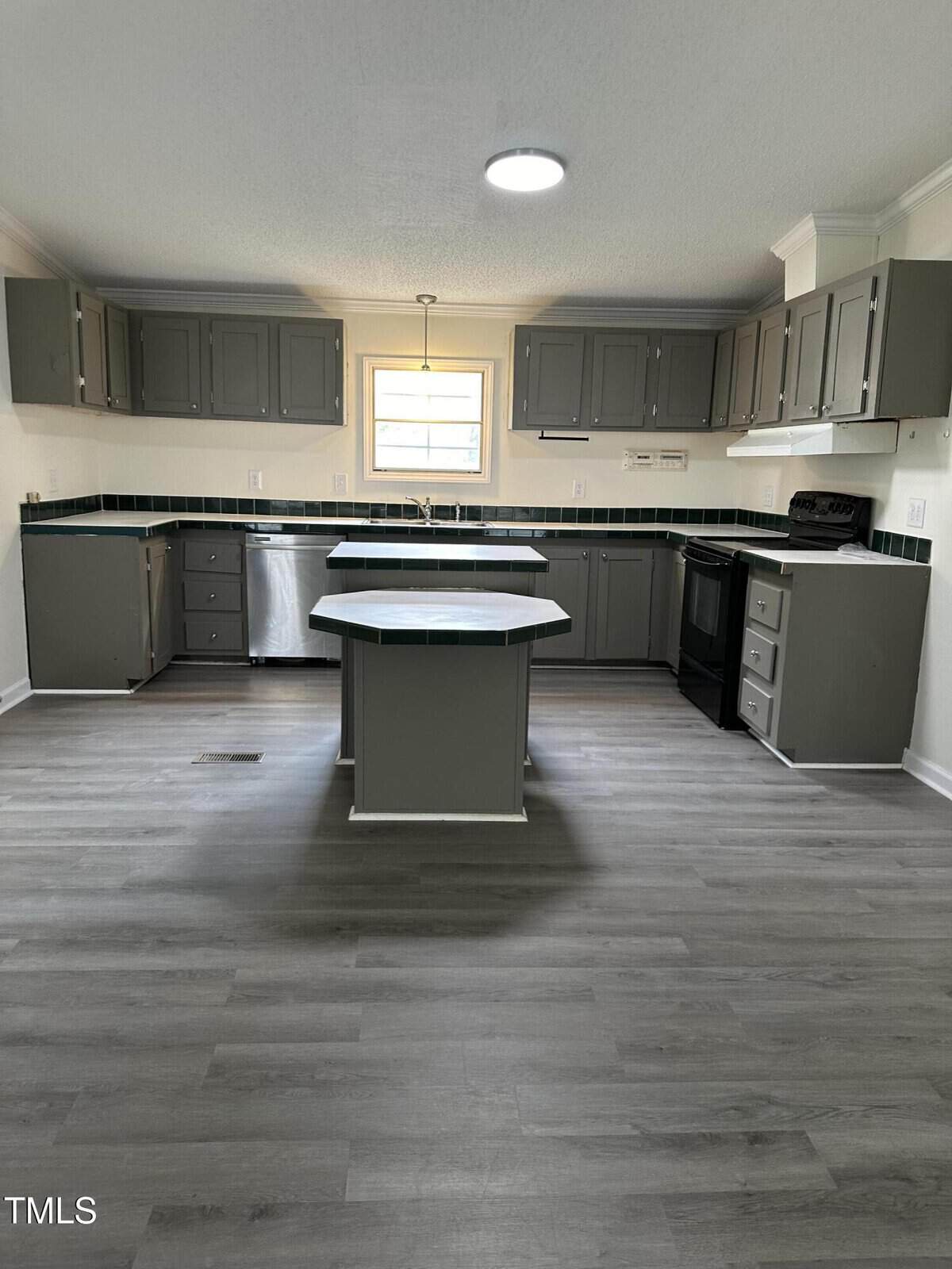 A modern kitchen with gray cabinets and a central island. The room features stainless steel appliances, a large window above the sink, and wood-look flooring. The backsplash is dark, contrasting with the light walls.