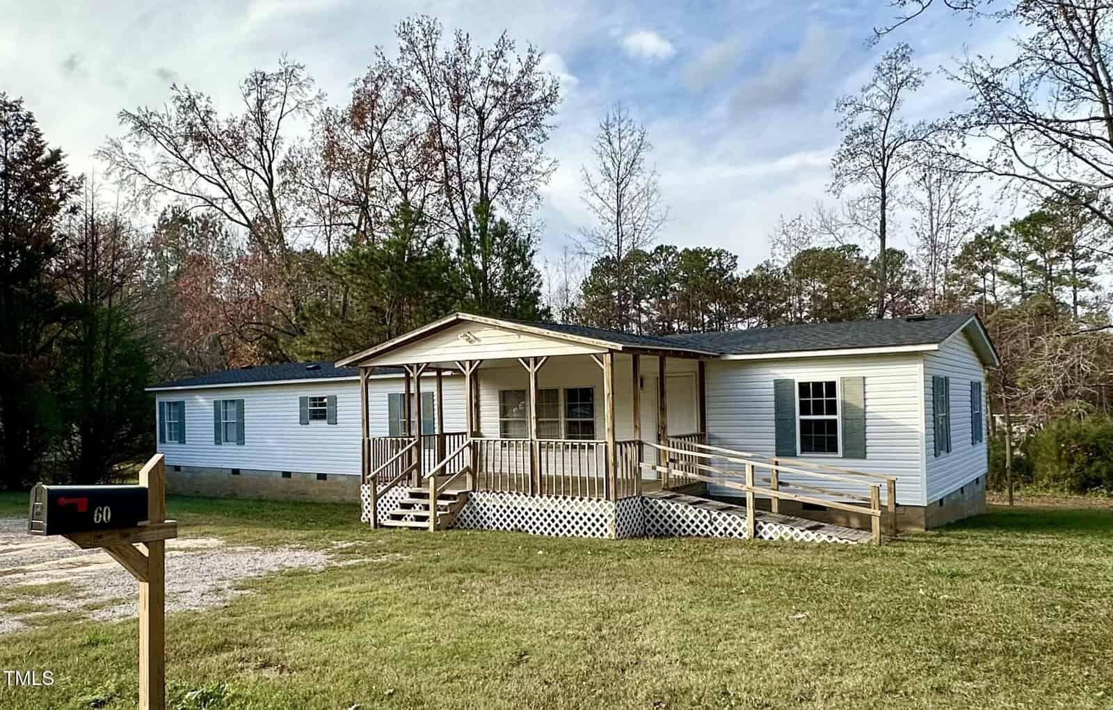 A white mobile home with a wooden ramp and porch sits on a grassy plot at 60 Casei Lane, Henderson, surrounded by trees. A mailbox bearing the number is visible on the left under the partly cloudy sky.