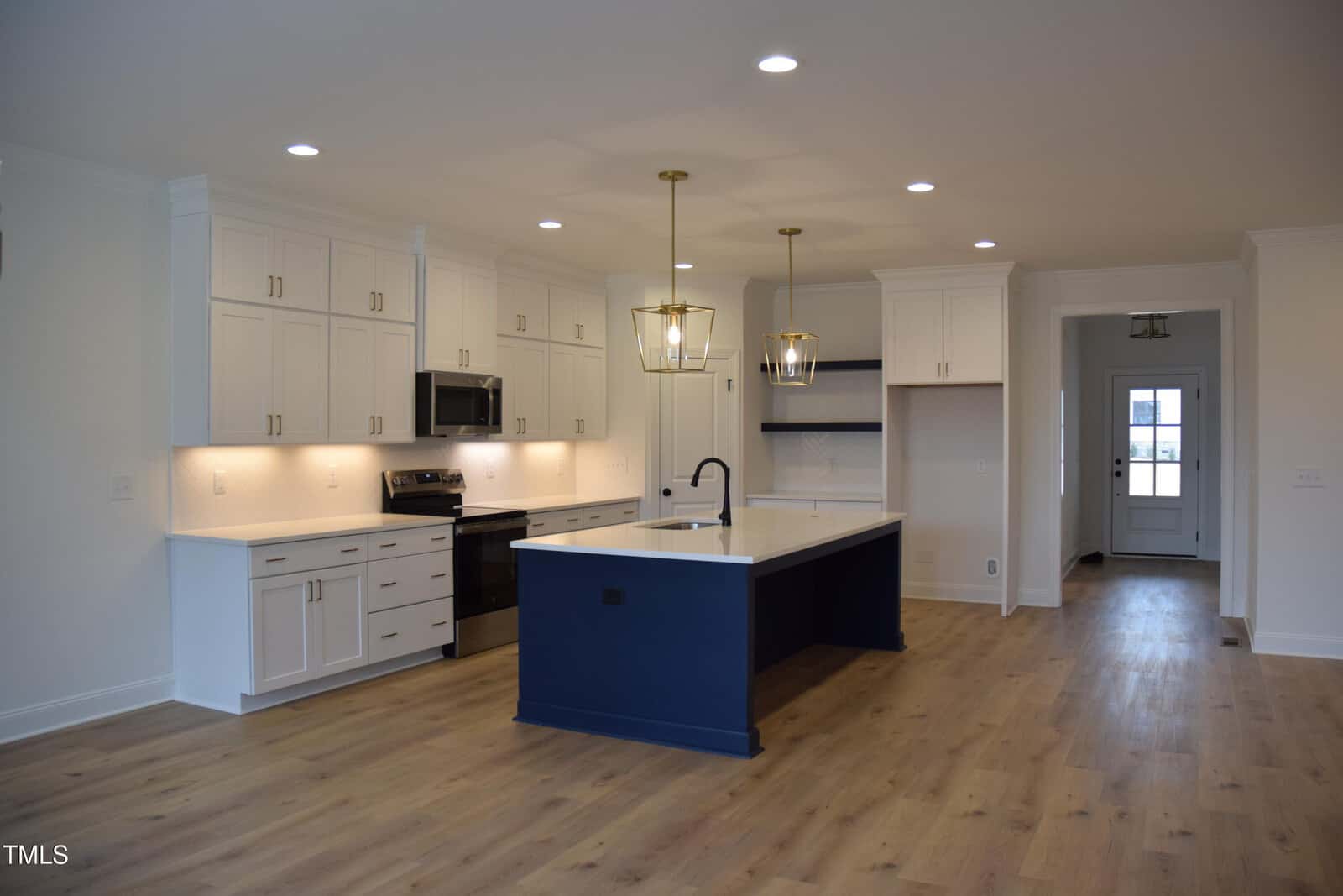 A modern kitchen with white cabinets, a large navy-blue island, and stainless steel appliances. Two pendant lights hang above the island. The room has wooden floors and recessed ceiling lights. A hallway leads to a door in the background.