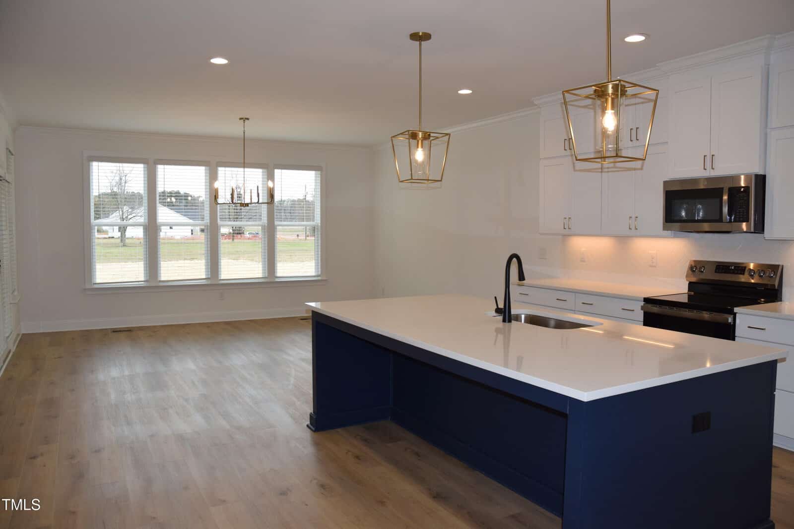 A modern kitchen with a large navy blue island featuring a white countertop and sink. Pendant lights hang above. White cabinets and a stainless steel stove and microwave are in the background. Wood flooring extends into a dining area with large windows.