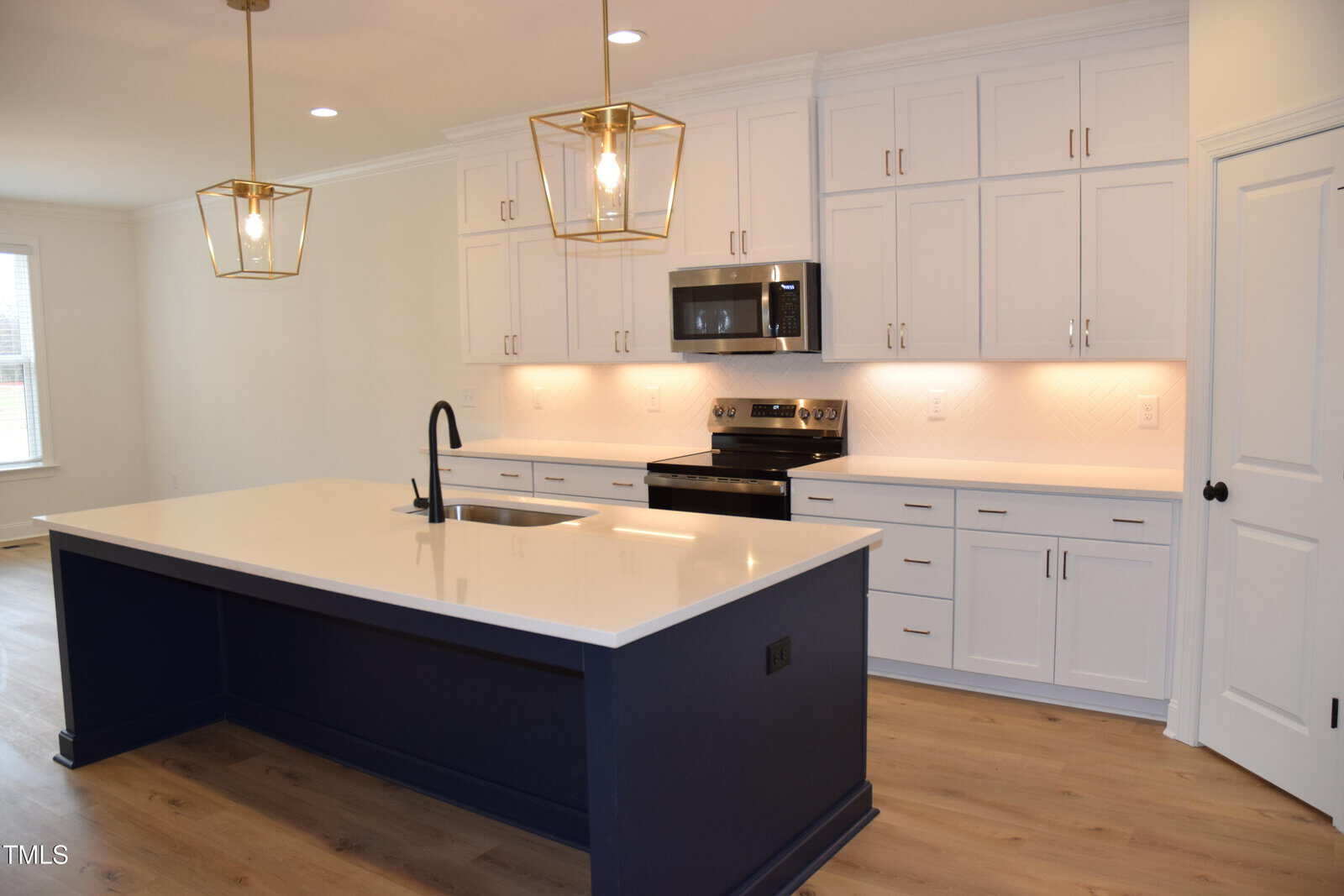 Modern kitchen with white cabinets, black appliances, and a large navy-blue island. Light wood flooring and brass pendant lights add warmth. Ample countertop space and recessed lighting provide a bright, clean look.
