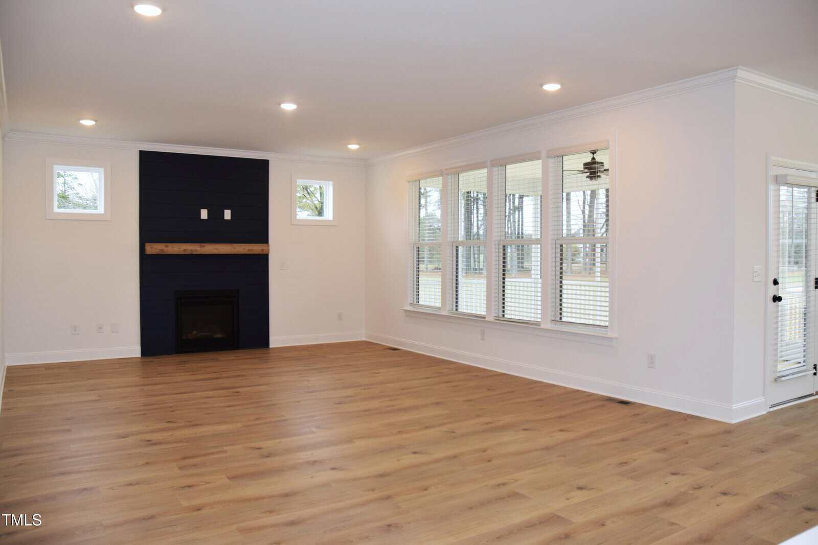 Spacious, empty room with light wood flooring, white walls, and recessed ceiling lights. Features a dark accent wall with a fireplace and wooden mantle. Large windows on the right offer abundant natural light.