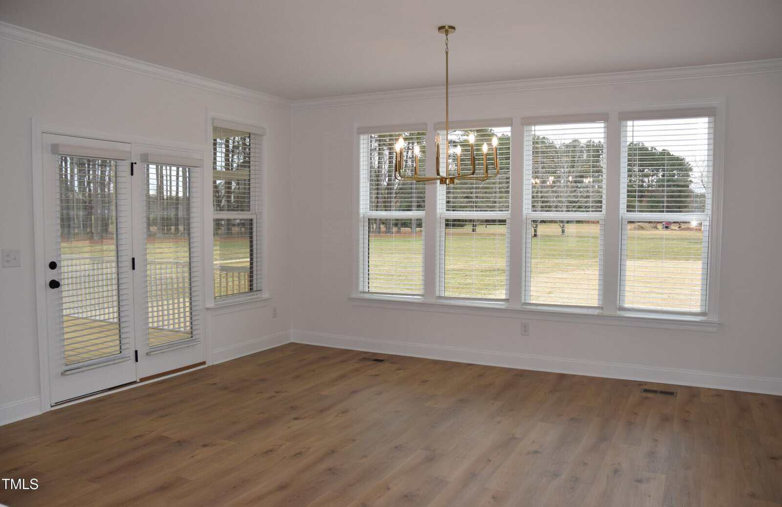 A bright, empty room with hardwood floors and white walls. Large windows with blinds provide natural light and overlook a grassy field. A modern chandelier hangs from the ceiling. A double glass door is on one side.