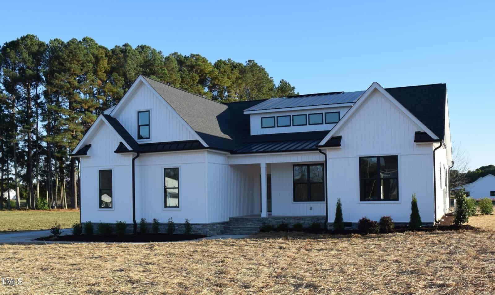 A modern, single-story white house with a dark roof, featuring multiple windows and a front porch. It's set on a newly landscaped lawn with freshly planted trees and shrubs, surrounded by a backdrop of tall trees under a clear blue sky.
