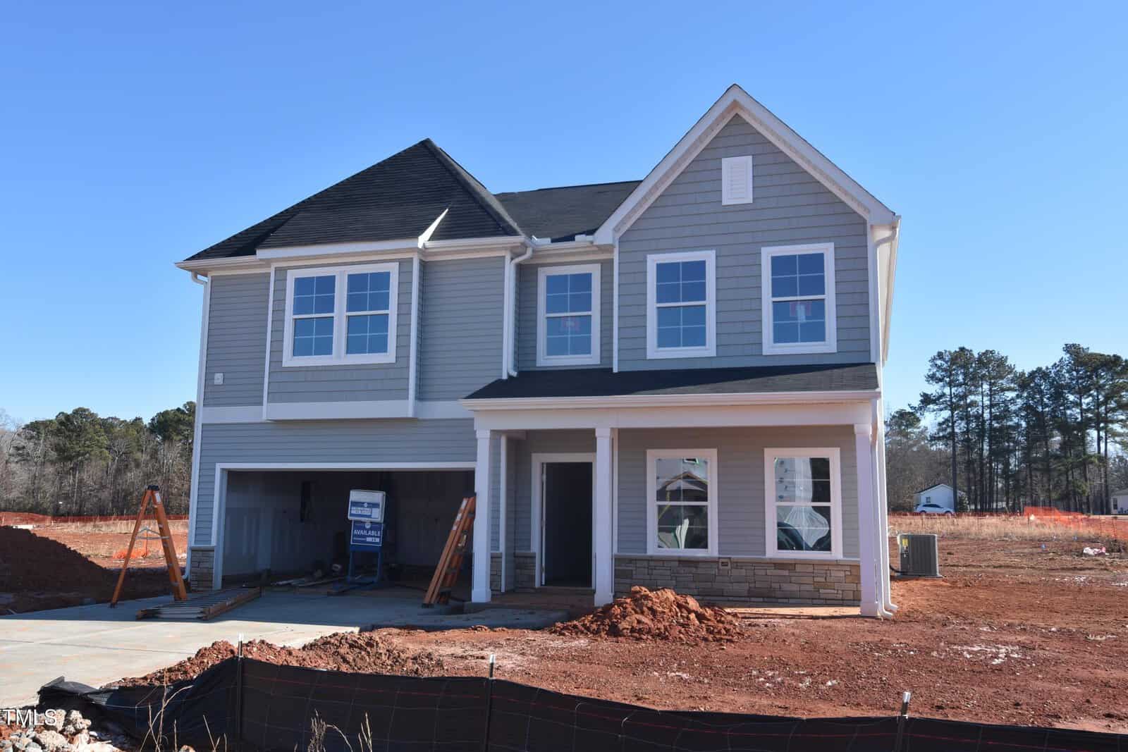 A two-story unfinished house with gray siding and white trim, featuring a covered front porch, double garage, construction materials on site, and red dirt piles in the yard against a clear blue sky.