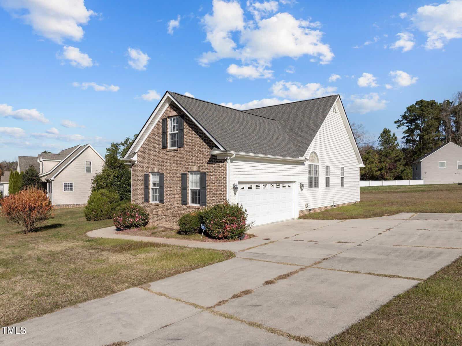 A single-story brick and siding house with a gabled roof sits on a spacious lawn. It features a two-car garage and a wide driveway. The sky is clear with scattered clouds, and neighboring houses are visible in the background.