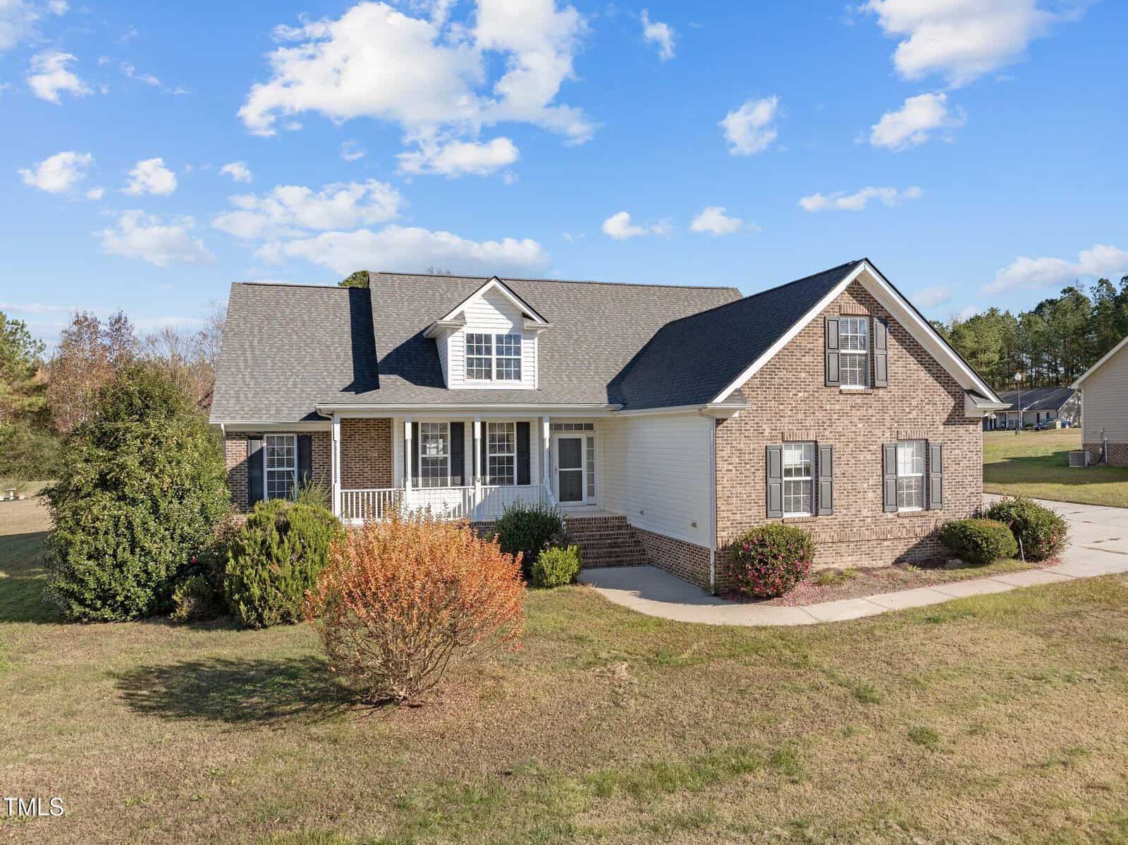 A large brick and white panel house with a gray roof, featuring front steps and a small porch. It's surrounded by grass, bushes, and trees, under a blue sky with scattered clouds.