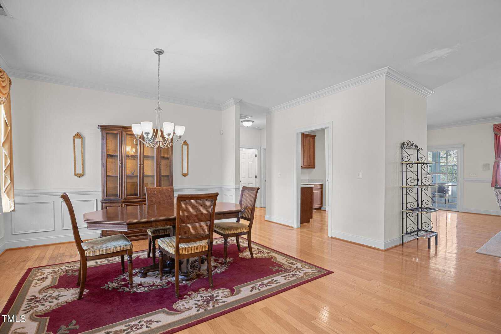 A dining room with a wooden table and four chairs on a red patterned rug. A chandelier hangs above the table. There's a wooden hutch against the wall and an open doorway leading to a hallway. Light-colored walls and hardwood floors complete the scene.