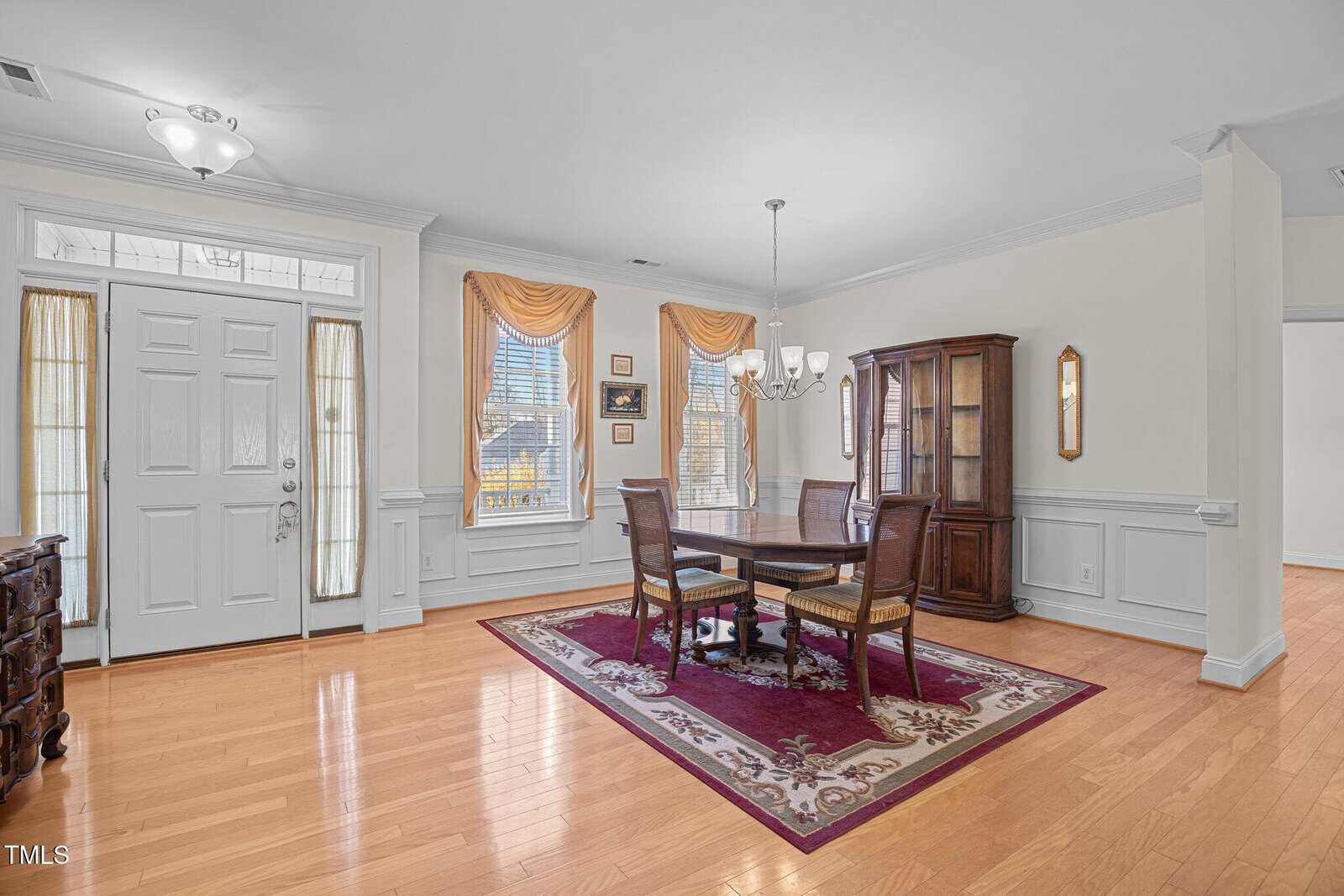 Bright dining area with wooden flooring, featuring a rectangular table with four chairs on a patterned rug. A chandelier hangs above the table. Large windows with drapes let in natural light. A wooden cabinet stands in the corner.