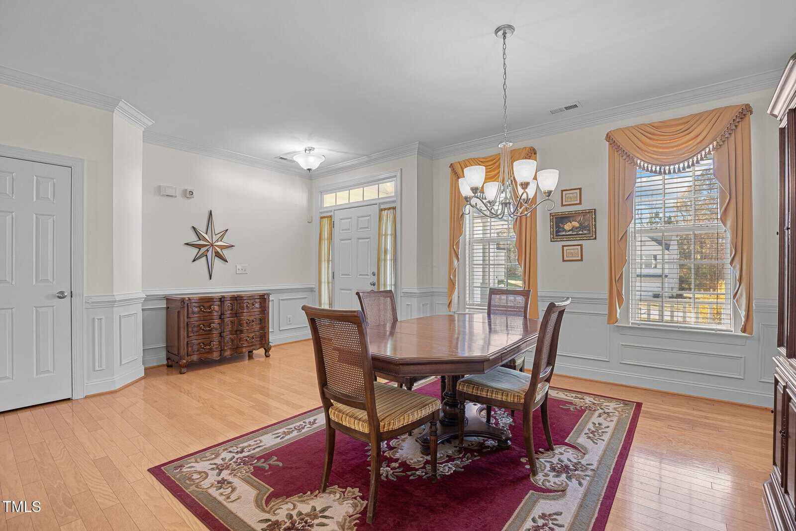 A dining room with a wooden table and six chairs on a red patterned rug. A chandelier hangs above. Large windows with drapes let in natural light. Walls are decorated with molding and a star-shaped wall piece. A sideboard is near the entrance.
