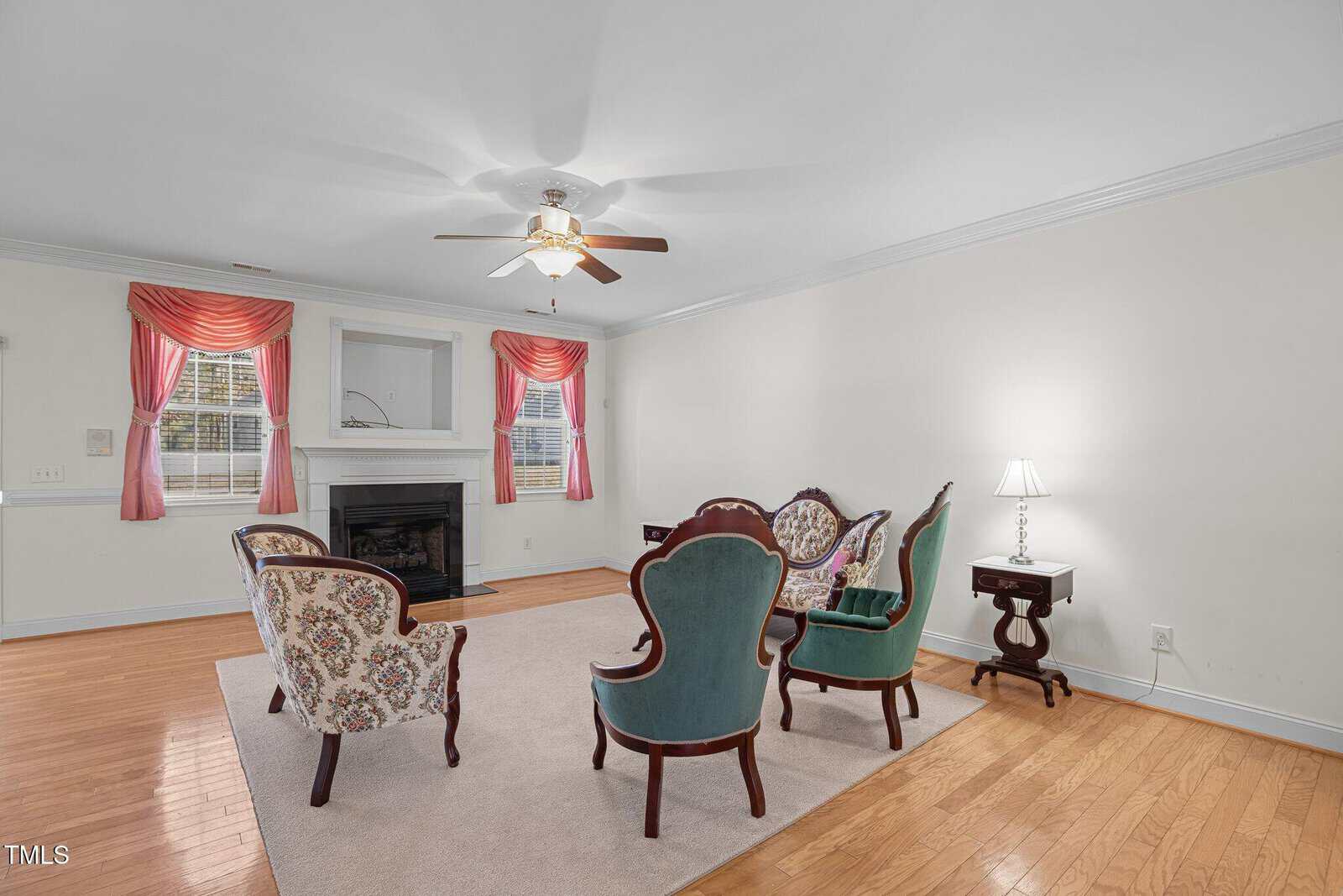 A living room with wooden flooring features vintage-style armchairs and a small table arranged around a fireplace. Pink curtains adorn the windows on a light-colored wall. A ceiling fan is centrally placed above the room.