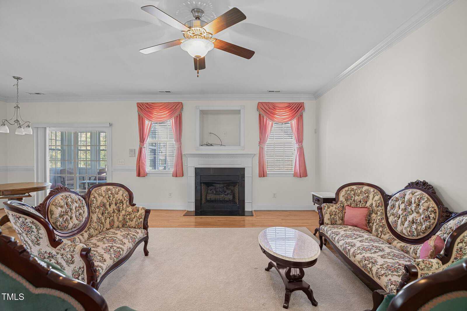A living room with vintage-style floral sofas and a wooden coffee table. A fireplace is set between two large windows with pink drapes. A ceiling fan with light hangs from the white ceiling. A dining area is visible in the background.