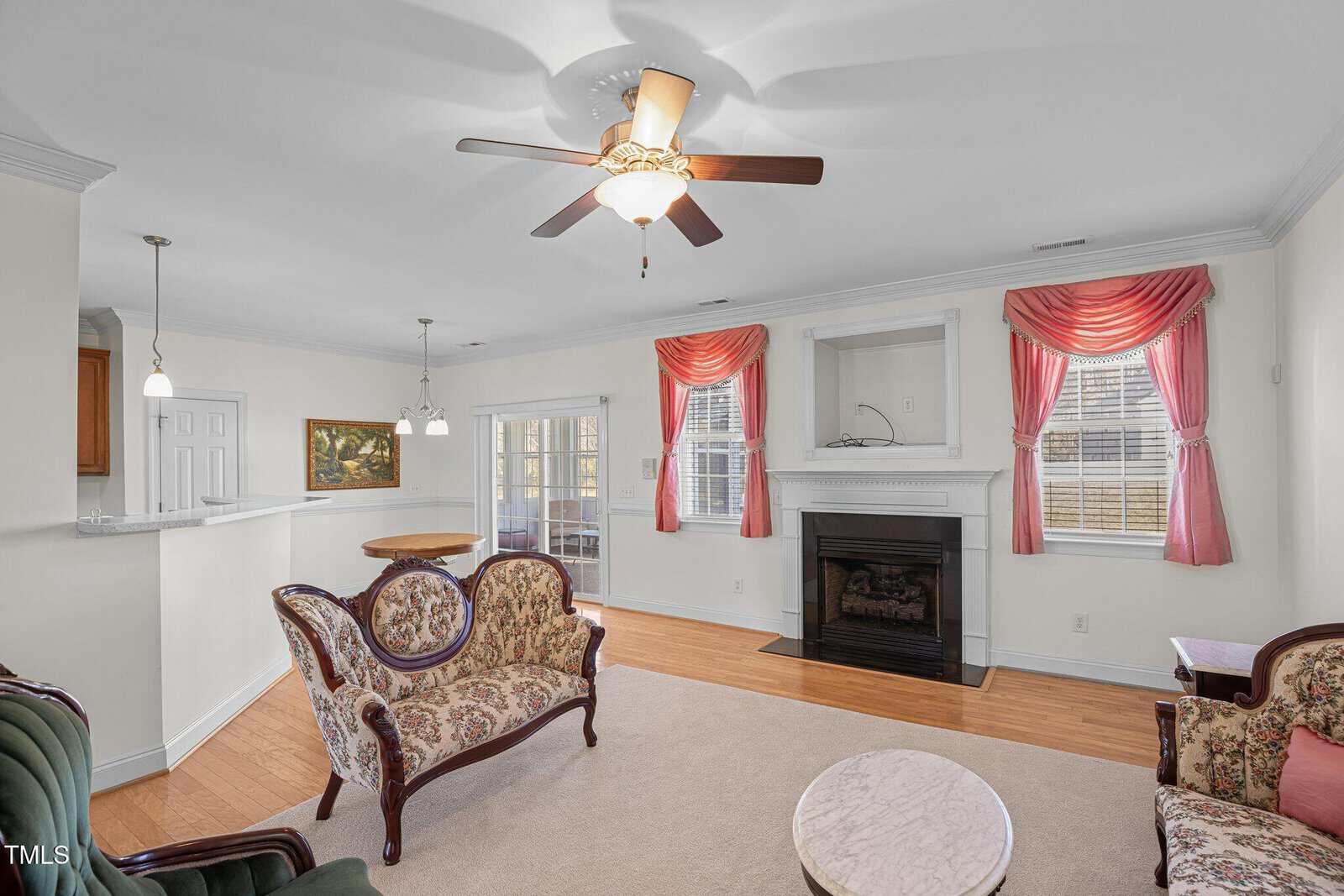 A bright living room with wooden floor, a ceiling fan, and a fireplace. The room features a vintage-style sofa set, pink curtains, and a mirror above the mantel. To the left, there's an open doorway leading to a kitchen area.