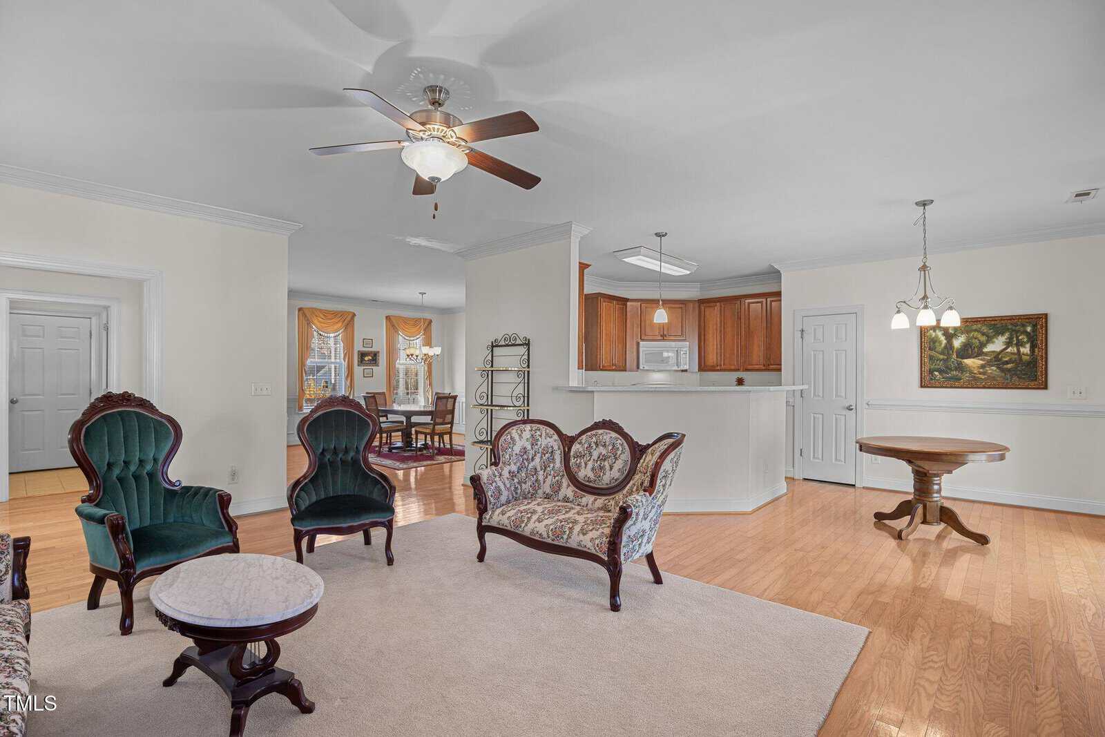 Spacious living area with wooden floors and a ceiling fan. Features vintage-style armchairs, a small sofa, and round tables with marble tops. To the right is a kitchen with wooden cabinets and a breakfast bar. Doorways lead to other rooms.