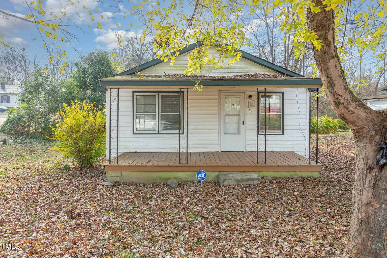 A small, single-story house at 413 Sycamore Street features white siding and a charming front porch. The yard is cloaked in fallen leaves, with a tree boasting vibrant yellow foliage to the right. An ADT security sign rests on the ground near the porch, ensuring peace of mind in Oxford.