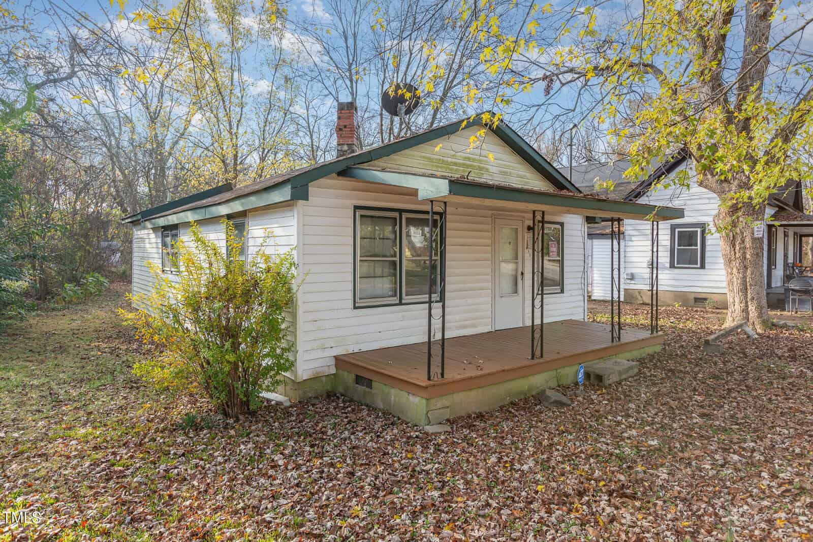 A small, white single-story house with a green roof and a covered front porch. The yard is covered in fallen leaves, and trees with yellowing leaves surround the house. Another similar house is partially visible on the right.