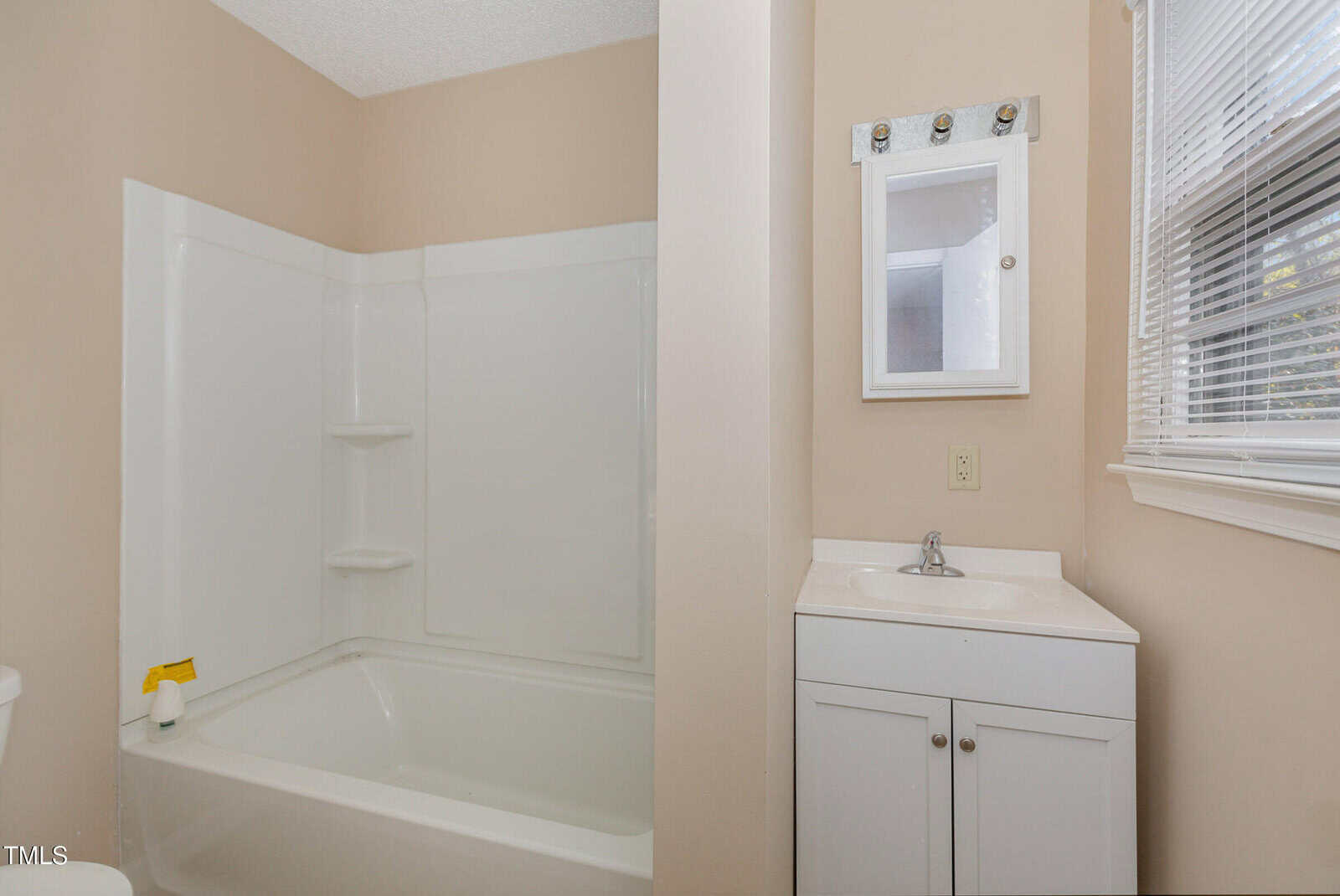 A bathroom with a white bathtub and shower combo on the left, featuring built-in shelves. On the right, there is a small white vanity with a mirror above it. A window with blinds is partially visible. The walls are painted beige.