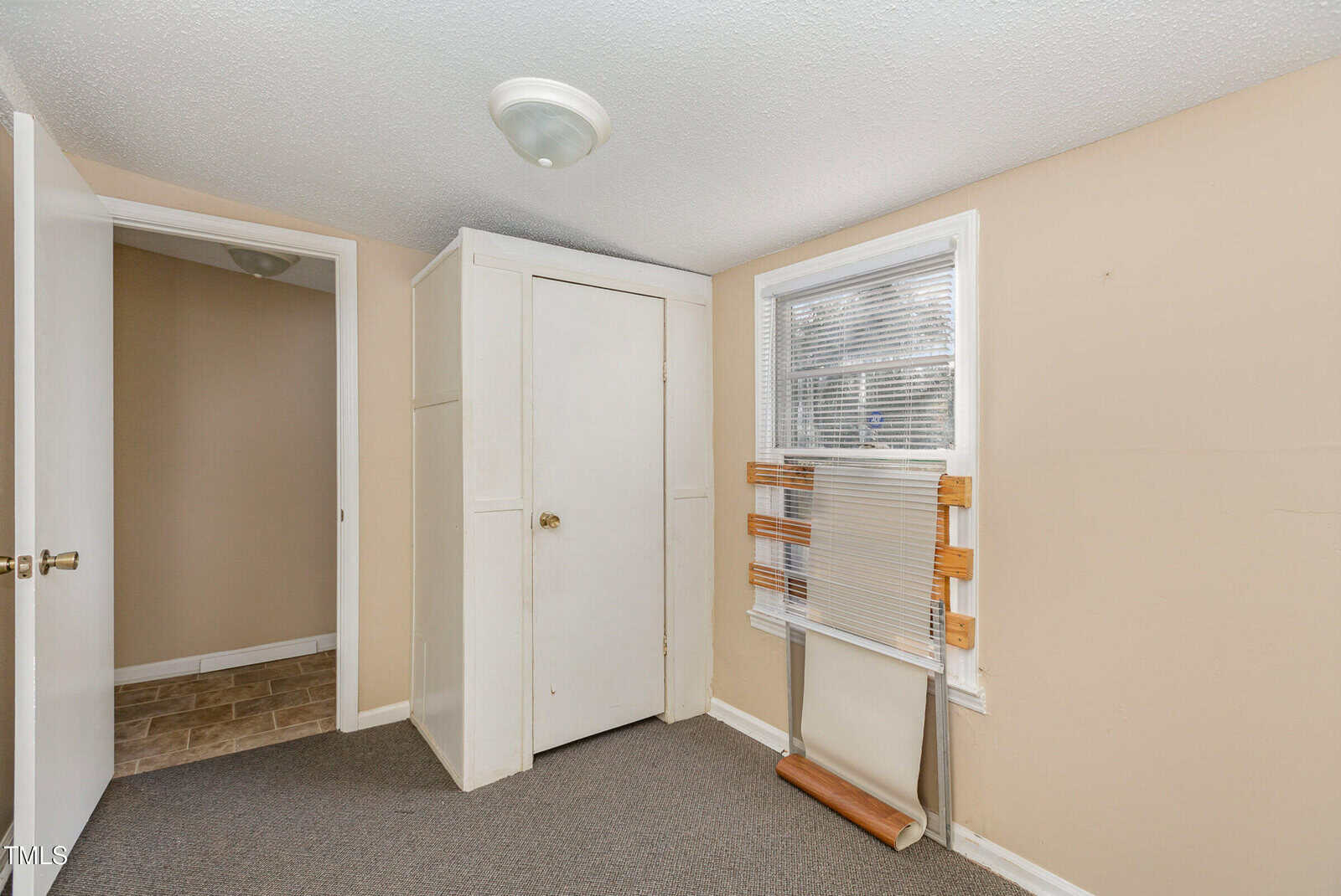 A small, empty room with beige walls and a gray carpet. It features a closed white door, a built-in white closet, and a window with a partially collapsed blind. A ceiling light fixture is present, and the doorway leads to a hallway with tiled flooring.