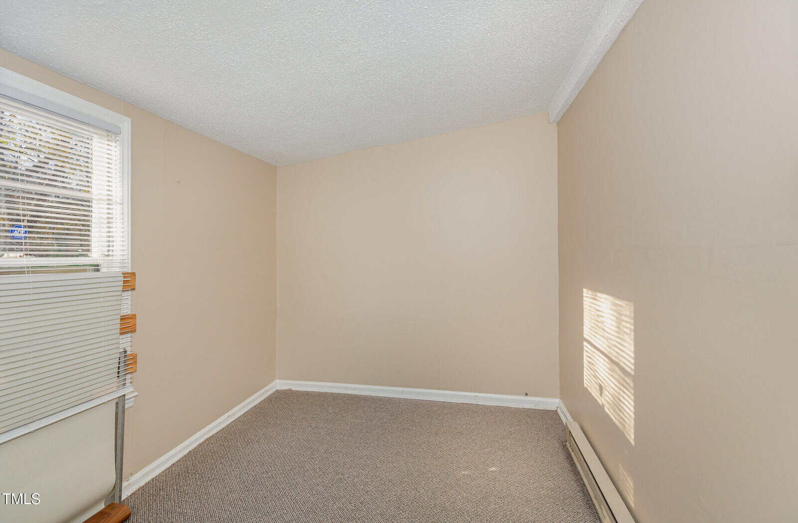 A small, empty room with beige walls and a beige carpet. There is a window on the left with closed blinds partially open, allowing some light in. The room appears clean and has a white baseboard along the bottom of the walls.