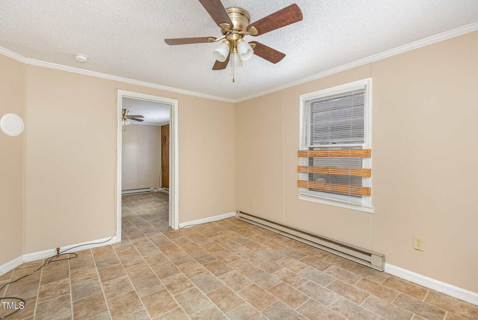 An empty room with beige walls and a tiled floor. A white ceiling features a brown and white ceiling fan with lights. There is a window with blinds on the right wall and an open doorway leading to another room with similar flooring.