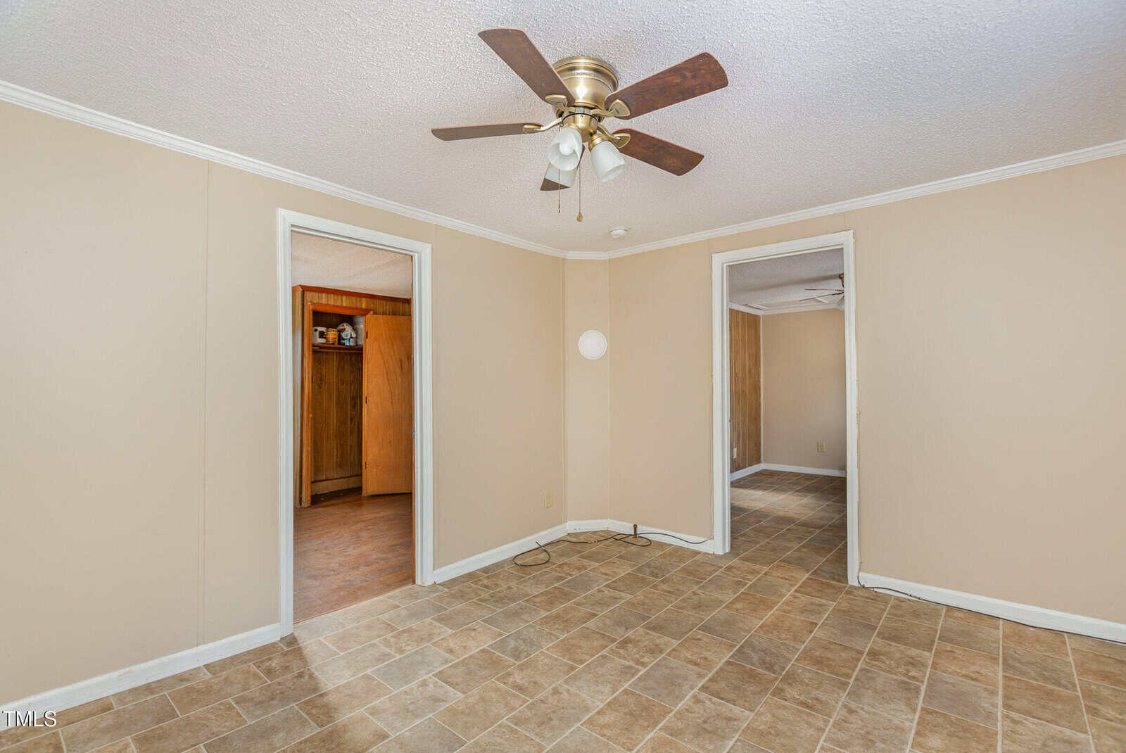 An empty room with beige walls and tiled flooring. Two open doorways lead to adjacent rooms. A ceiling fan with lights is centered above. The room is well-lit by natural light.