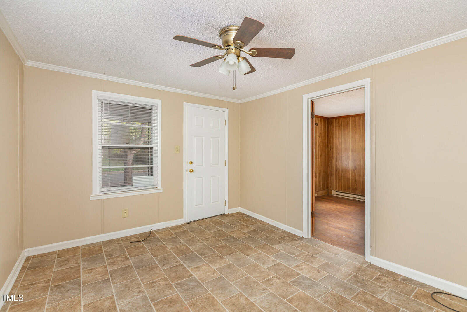 An empty room with beige walls, a tile floor, and a ceiling fan. A white door and a window are on one wall. An open doorway leads to a room with wood paneling.
