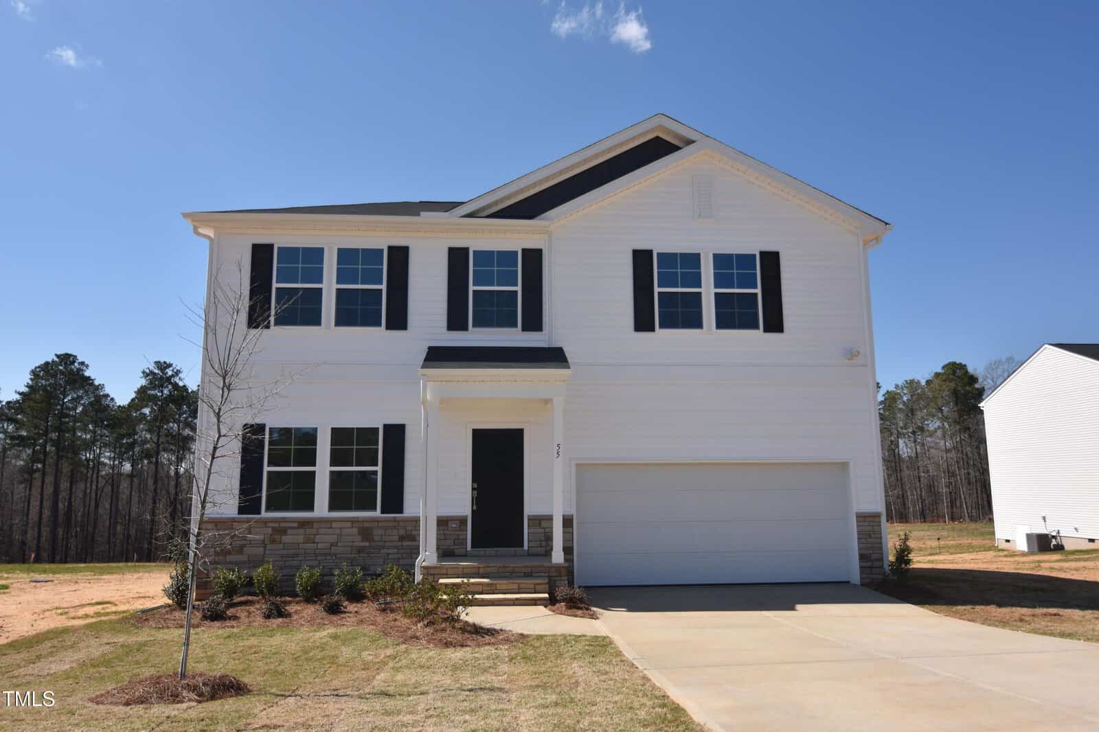 A two-story modern suburban house with white siding, black shutters, a double garage, and a small covered front porch. The yard is partially landscaped, and the sky is clear with no visible clouds.