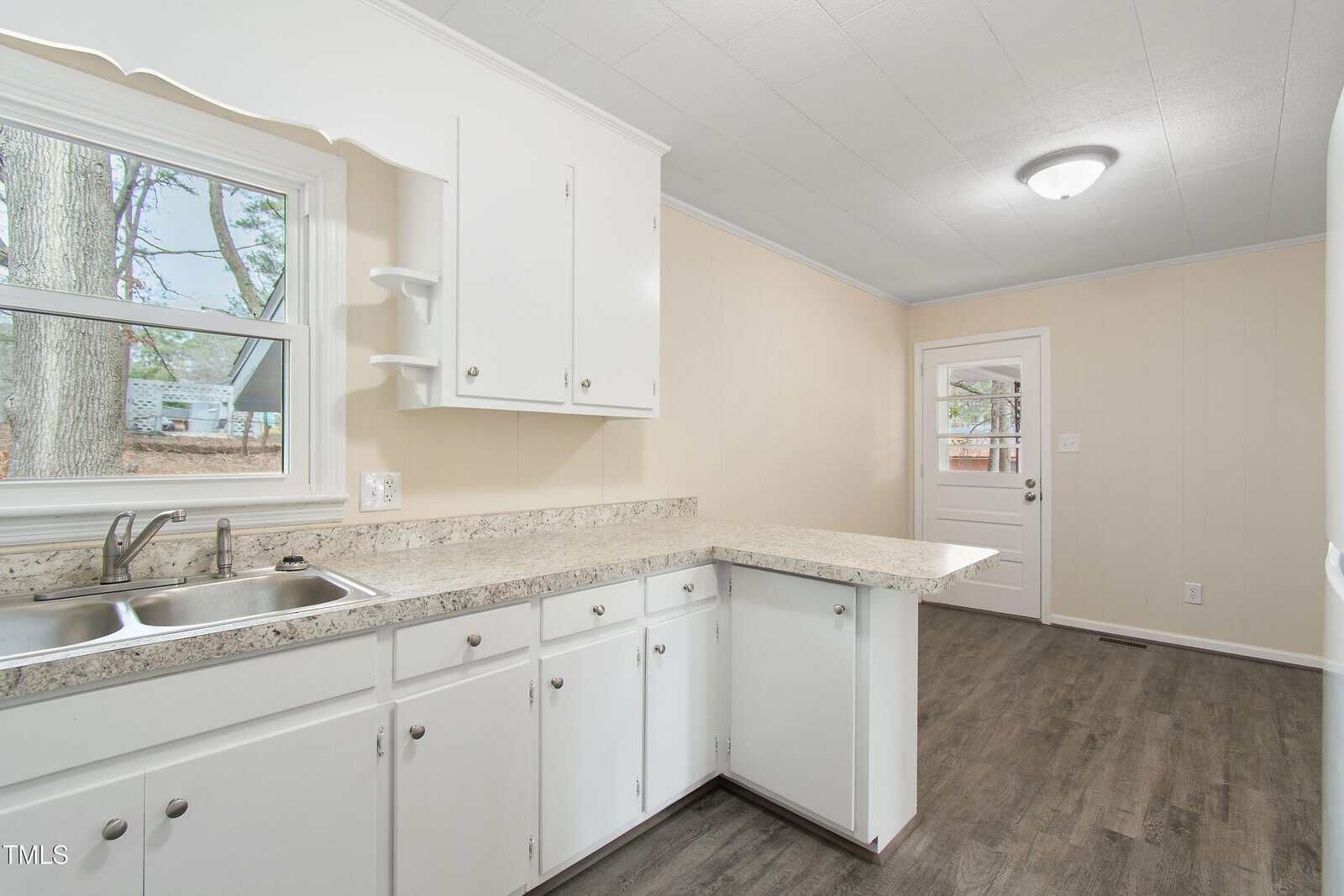 A kitchen with white cabinets, a dual sink, and a light granite countertop. The floor is dark wood, and there is a window above the sink. A door with a window is in the background, leading to another room.