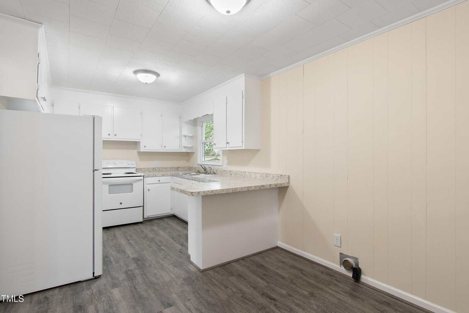 A small, empty kitchen featuring white cabinets, a white refrigerator, and a white stove. Light wood-patterned flooring contrasts with cream-colored walls. A single window above the sink provides natural light.