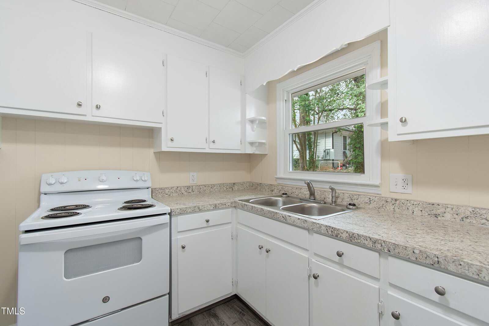 A small kitchen featuring white cabinets, a marble-effect countertop, a white electric stove with four burners, a double stainless steel sink, and a window above the sink. The floor is dark wood, and the walls are light beige.