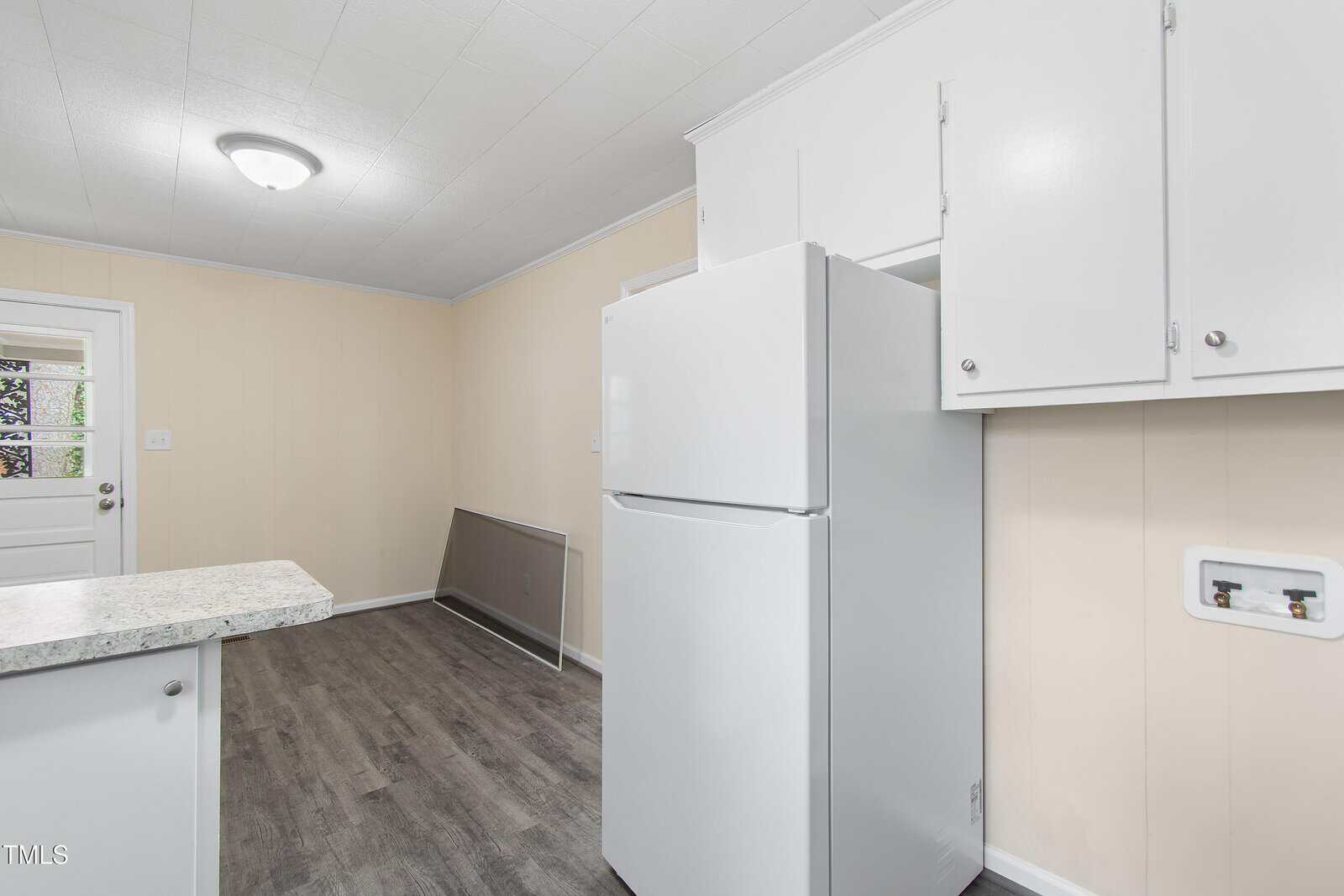 A kitchen with light-colored walls and dark flooring features a white refrigerator and white cabinetry. A small section of countertop is visible. A closed door with a window is on the left side of the room.