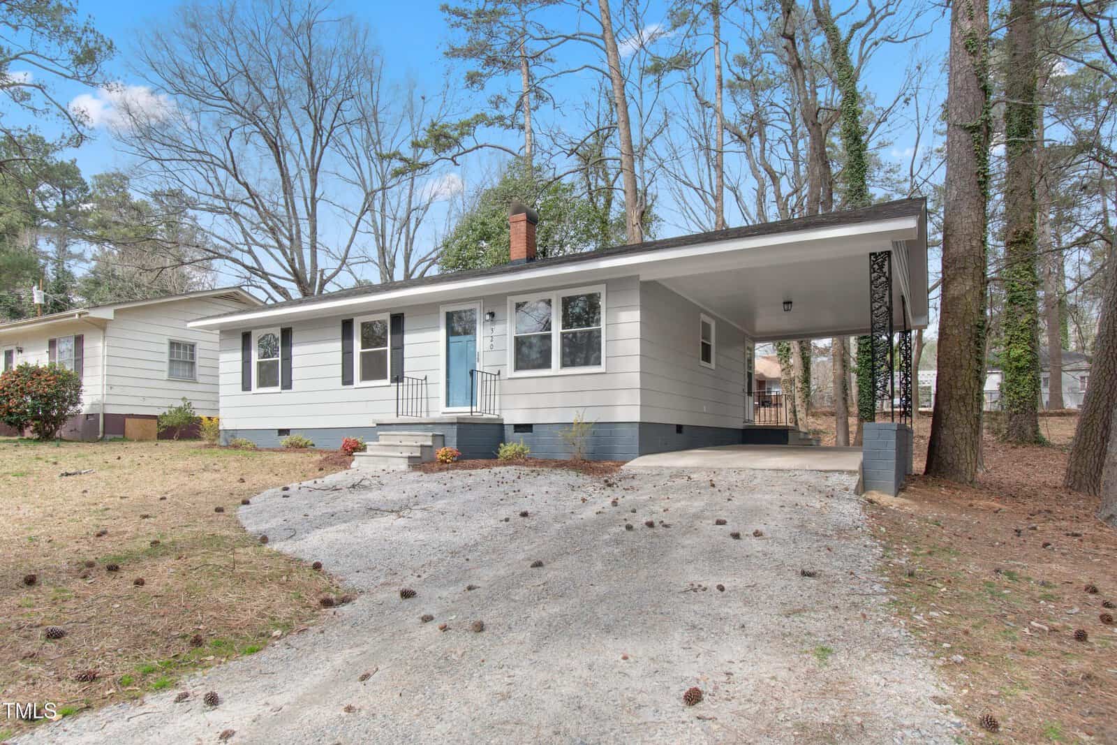 Single-story white house with a light blue door, a chimney, and a covered carport. Situated on a sloped driveway surrounded by trees and a patchy lawn. The sky is clear and blue.