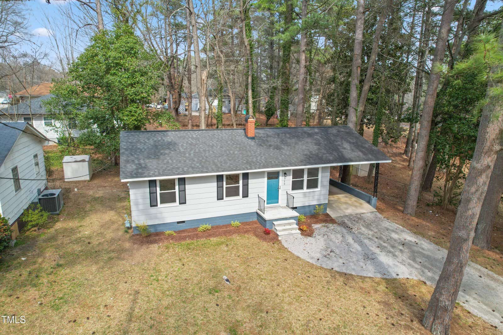 A small, single-story house with a gray exterior and a blue front door. It has a black shingled roof and a paved driveway. The house is surrounded by trees and a light brown grass lawn. Another house is partially visible on the left.