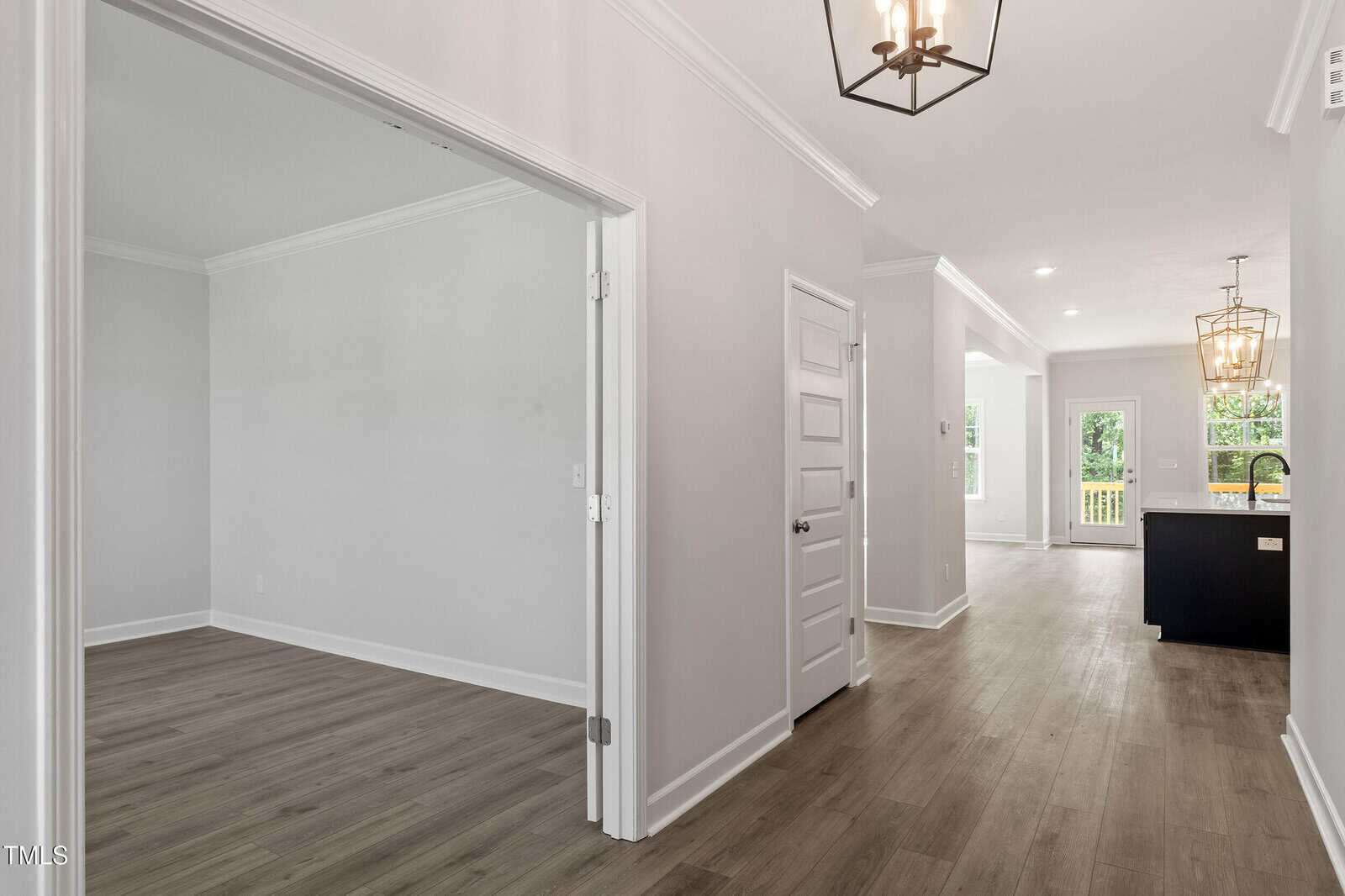 A hallway with wood floors leads to a kitchen and living area. Double doors open to a separate room on the left. The walls are light-colored with white trim, and there are ceiling lights and natural light from windows.
