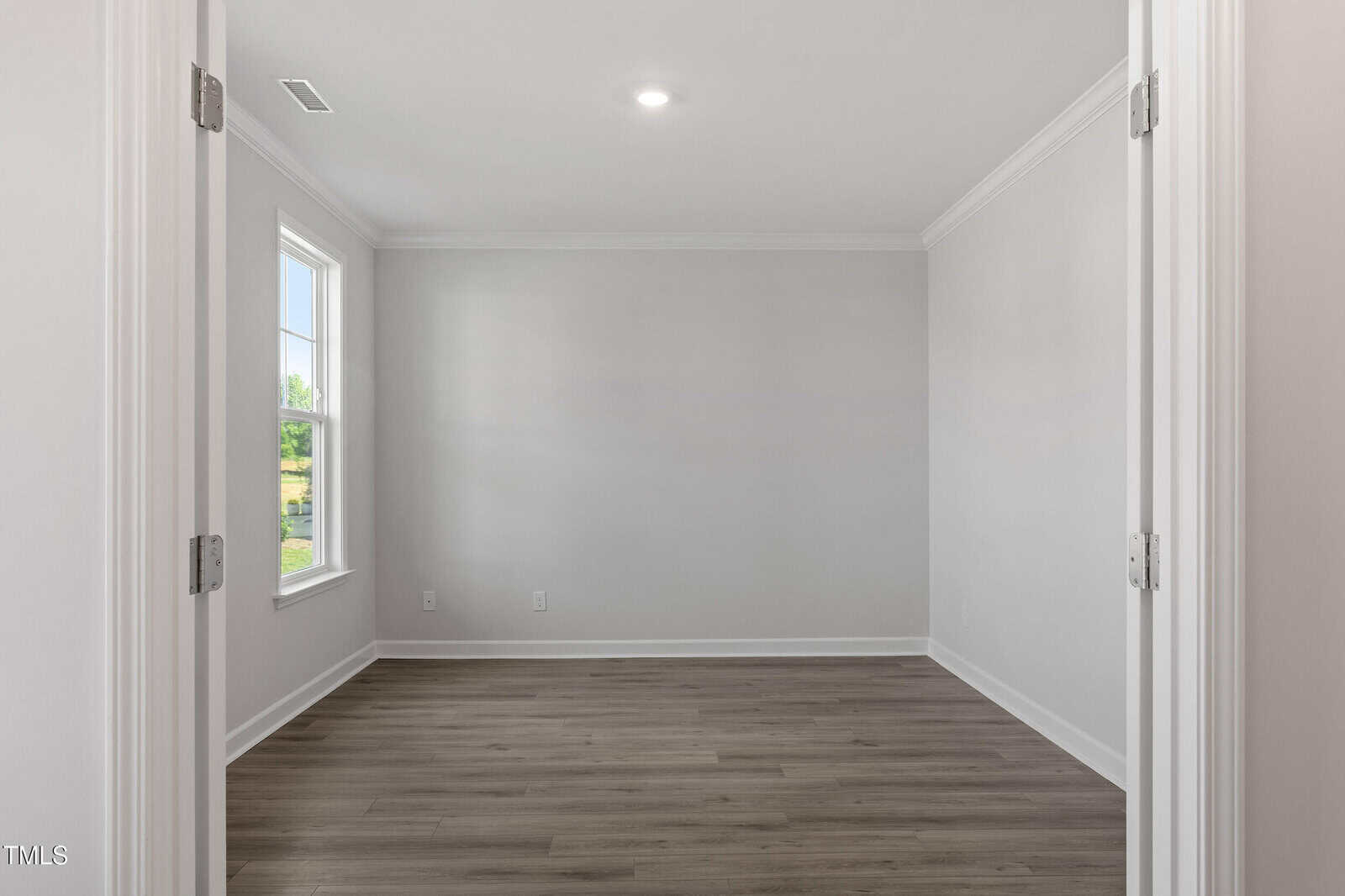 An empty room with light gray walls, white trim, wood-style flooring, a single window on the left wall, and a ceiling light. The room is viewed through double doors.