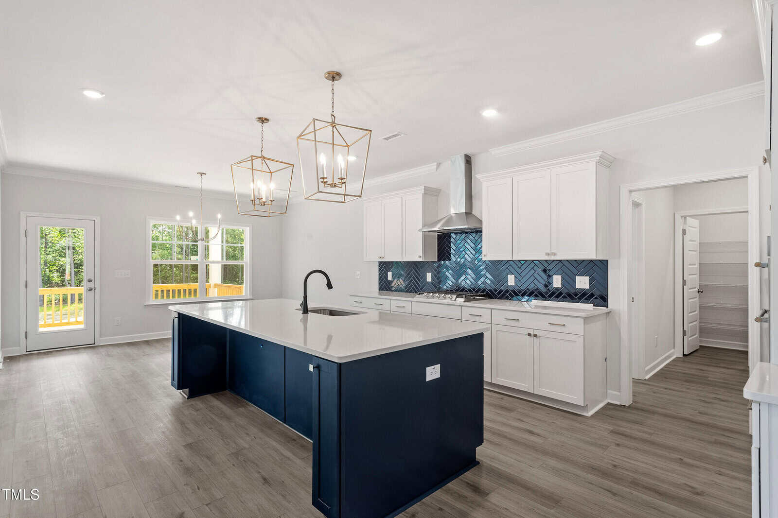 Modern kitchen with white cabinets, a large navy island, geometric gold pendant lights, blue tile backsplash, stainless steel appliances, wood flooring, and a dining area with large windows showing a view of trees and a deck.