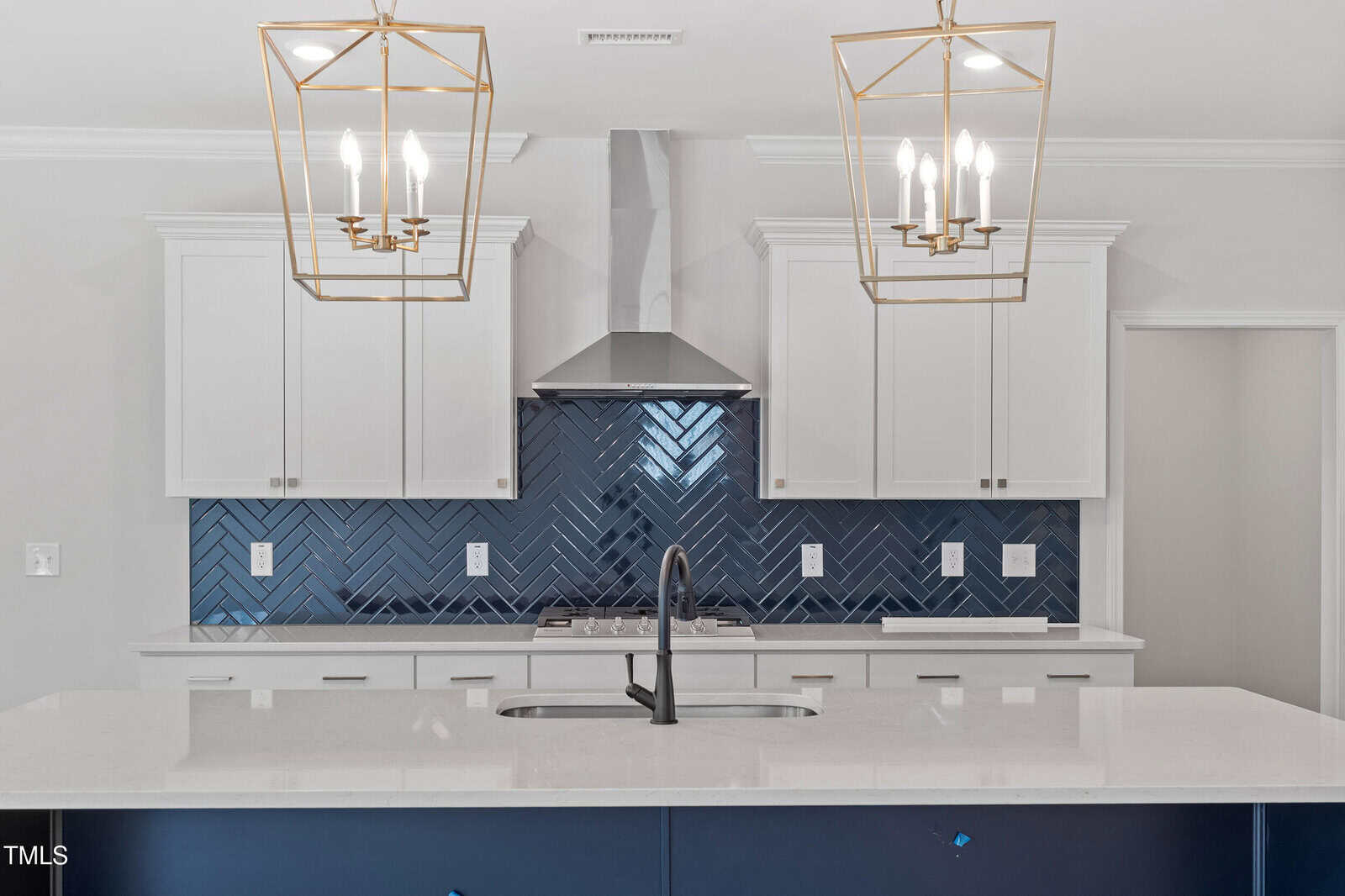 Modern kitchen with white cabinets, a stainless steel range hood, navy blue herringbone tile backsplash, quartz countertop island with a sink, and two gold geometric pendant lights hanging above the island.