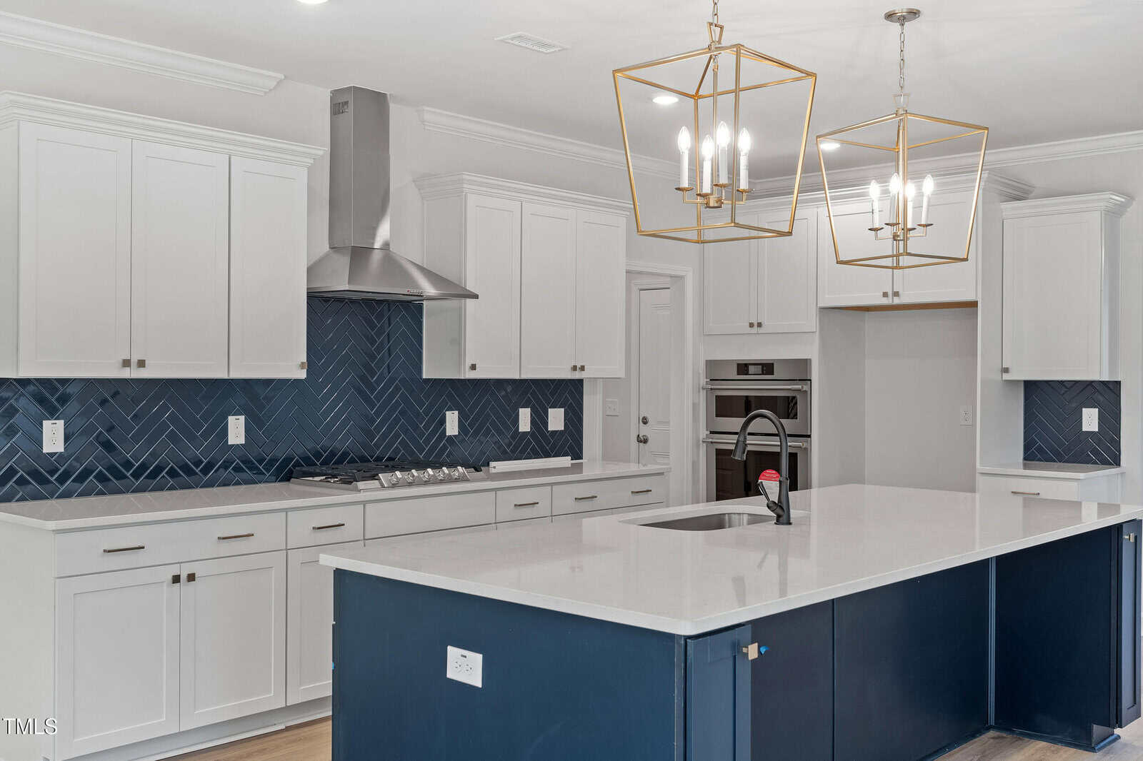 A modern kitchen with white cabinets, a large blue island, stainless steel appliances, two gold geometric pendant lights, and a blue herringbone tile backsplash. The countertops are white and uncluttered.