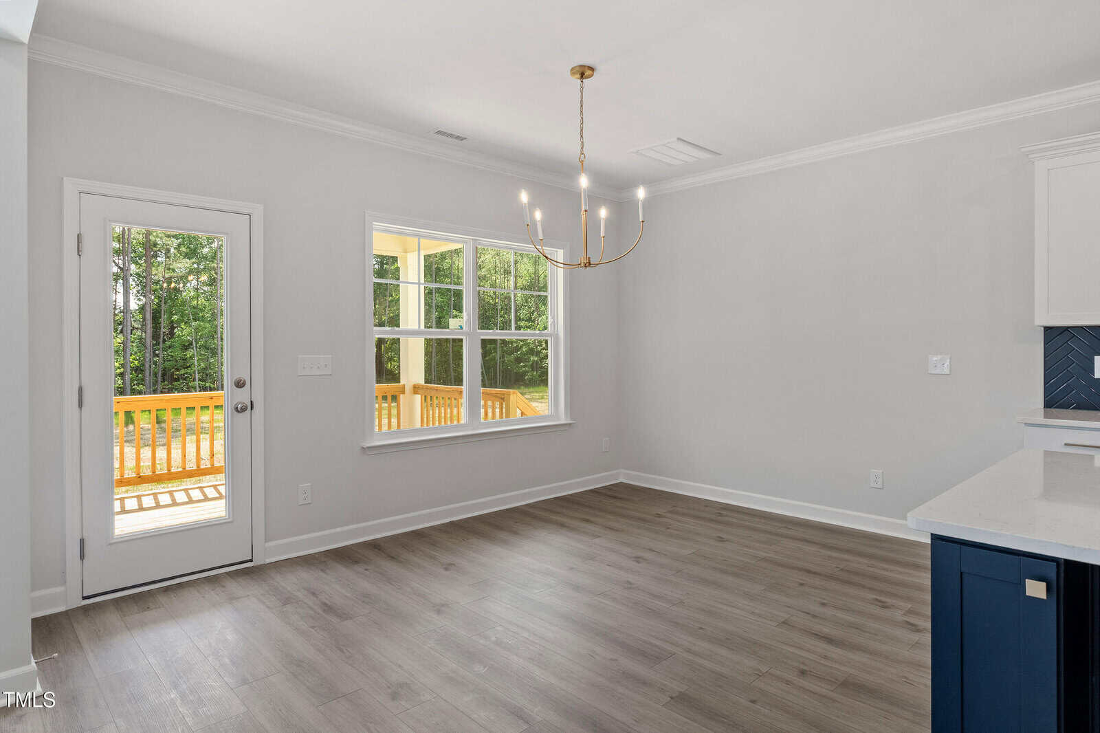 Empty dining area with light gray walls, wood-look flooring, a gold chandelier, and a window and glass door overlooking a wooden deck and trees outside. Part of a white and blue kitchen is visible on the right.